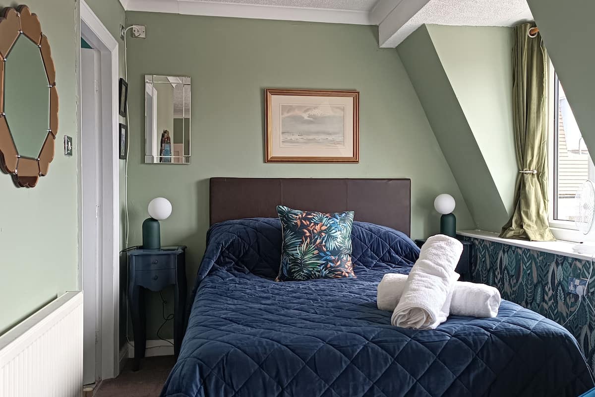 Un dormitorio con cama, mesita de noche, espejo, un cuadro enmarcado y ventana con cortinas.