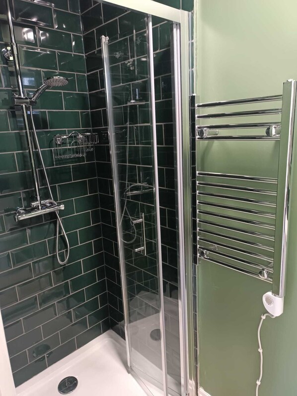 Ducha con cerramiento de vidrio, grifería cromada, bañera blanca y pared con azulejos verdes.