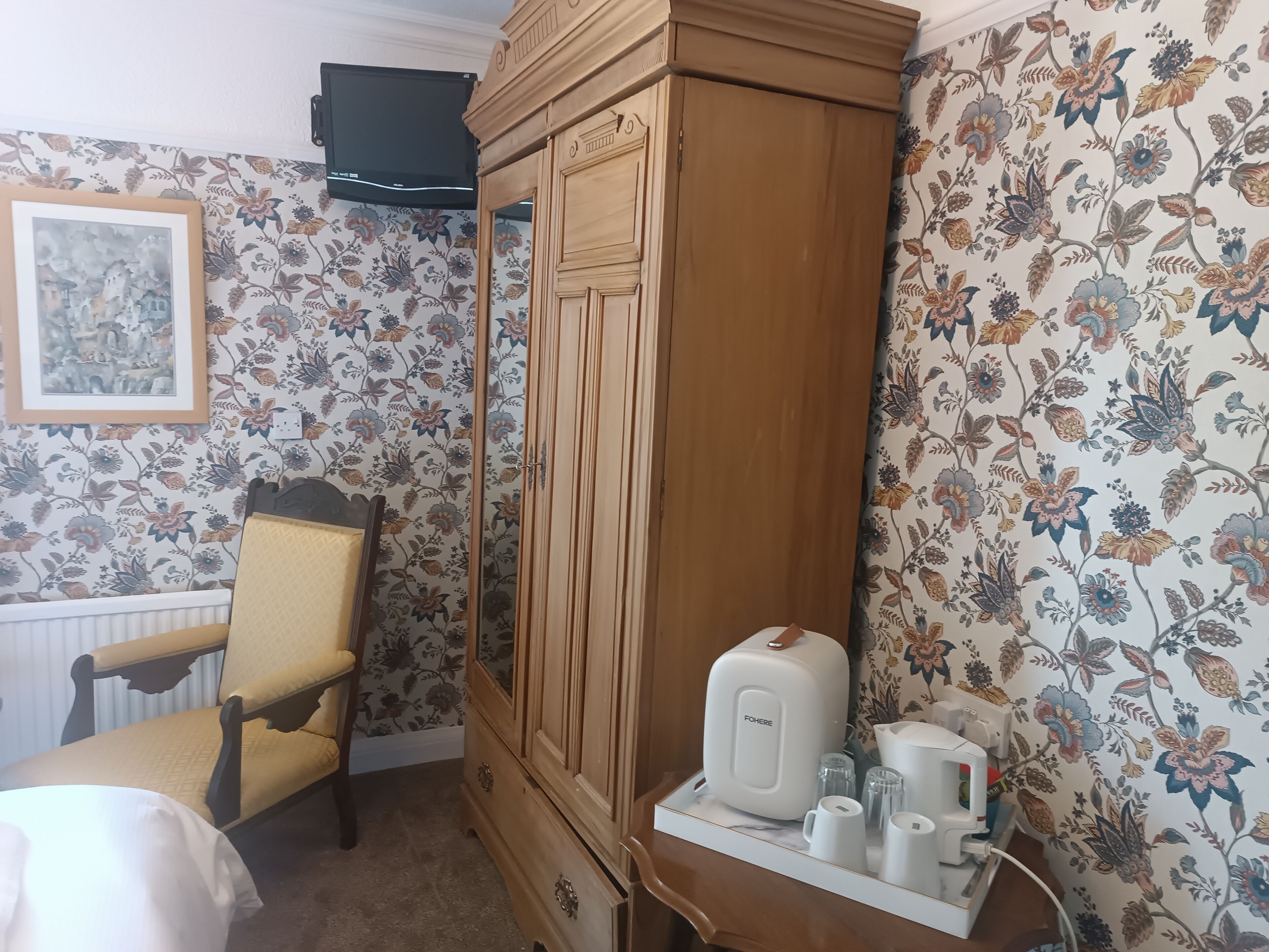 Habitació amb un armari de fusta, una cadira, un llit i una taula amb una cafetera i tasses.