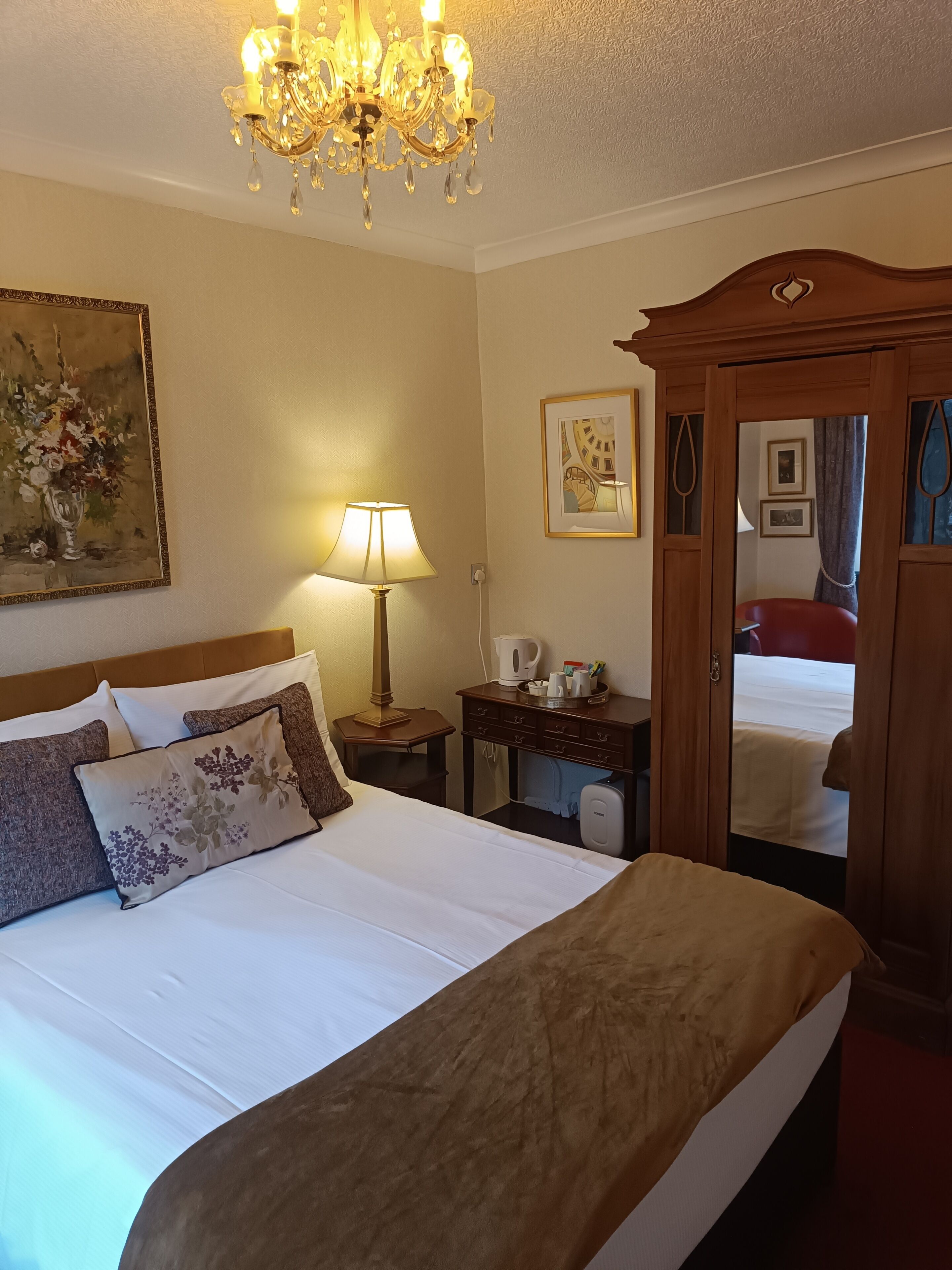 A hotel room with a bed, a nightstand, a lamp, a painting, a mirror, and a wooden wardrobe.