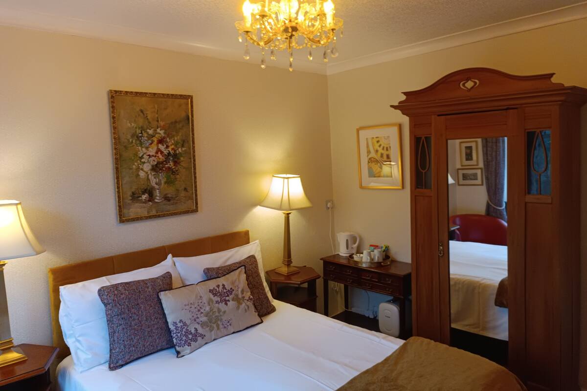 Quarto de hotel com cama, lustre, pintura, luminária, espelho e guarda-roupa de madeira.