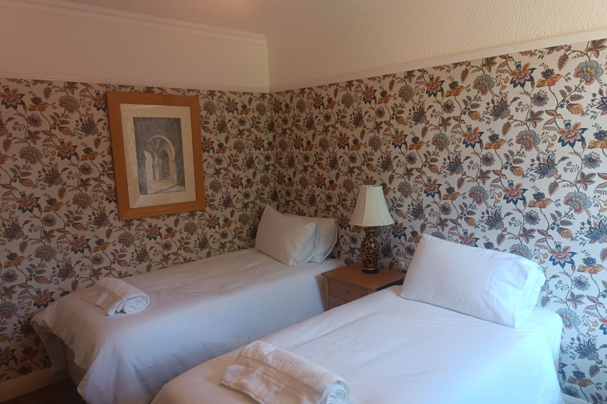 Quarto de hotel com duas camas, papel de parede floral, um quadro na parede e uma mesa de cabeceira com abajur.