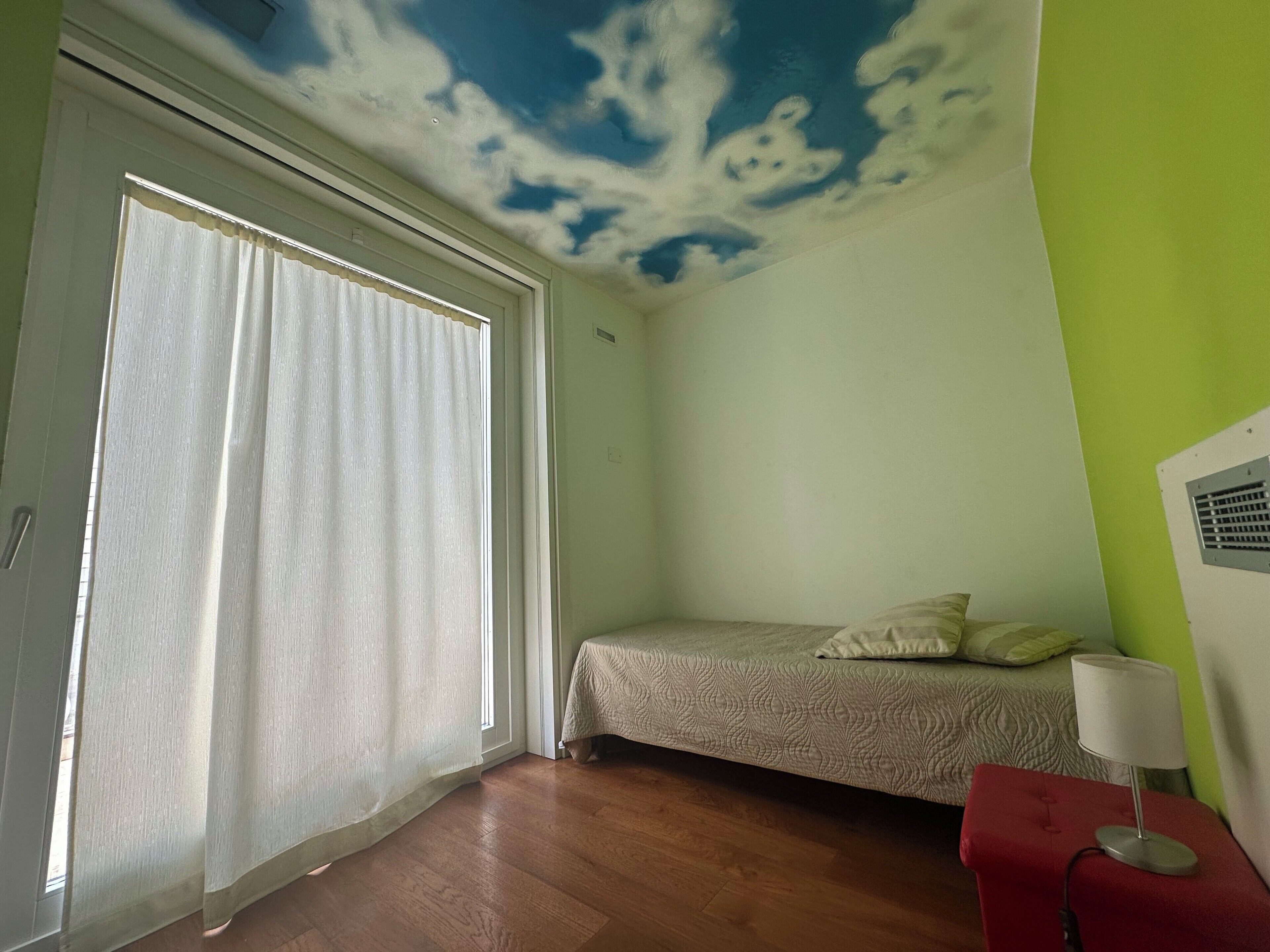 Une chambre à coucher avec un lit, une fenêtre avec des rideaux, un pouf rouge et un plafond décoré de motifs de nuages.