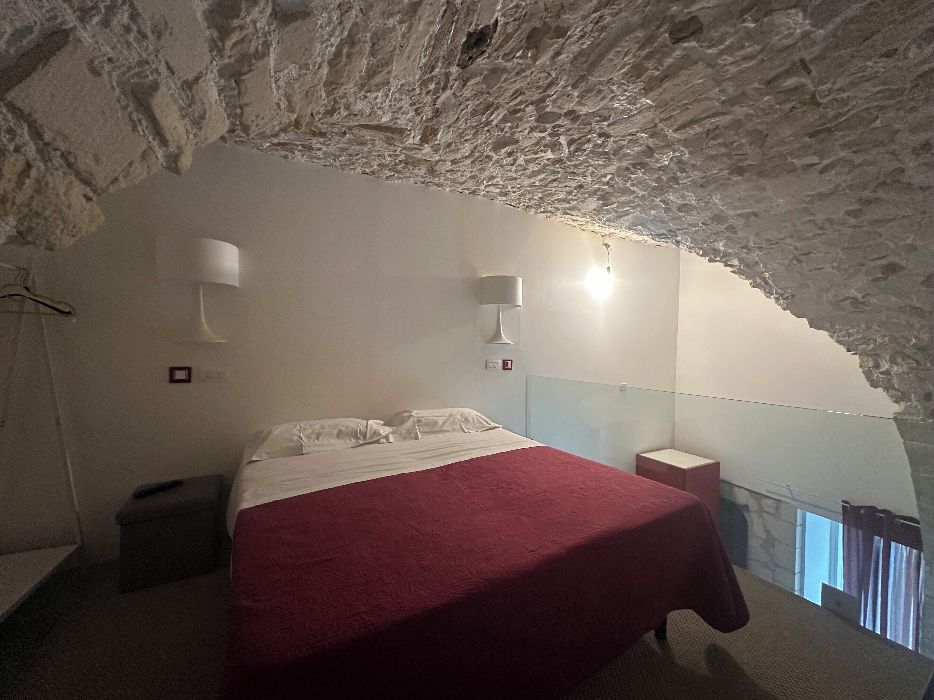 A bedroom with a bed, two wall-mounted lamps, a small bedside table, and a view through a glass partition.