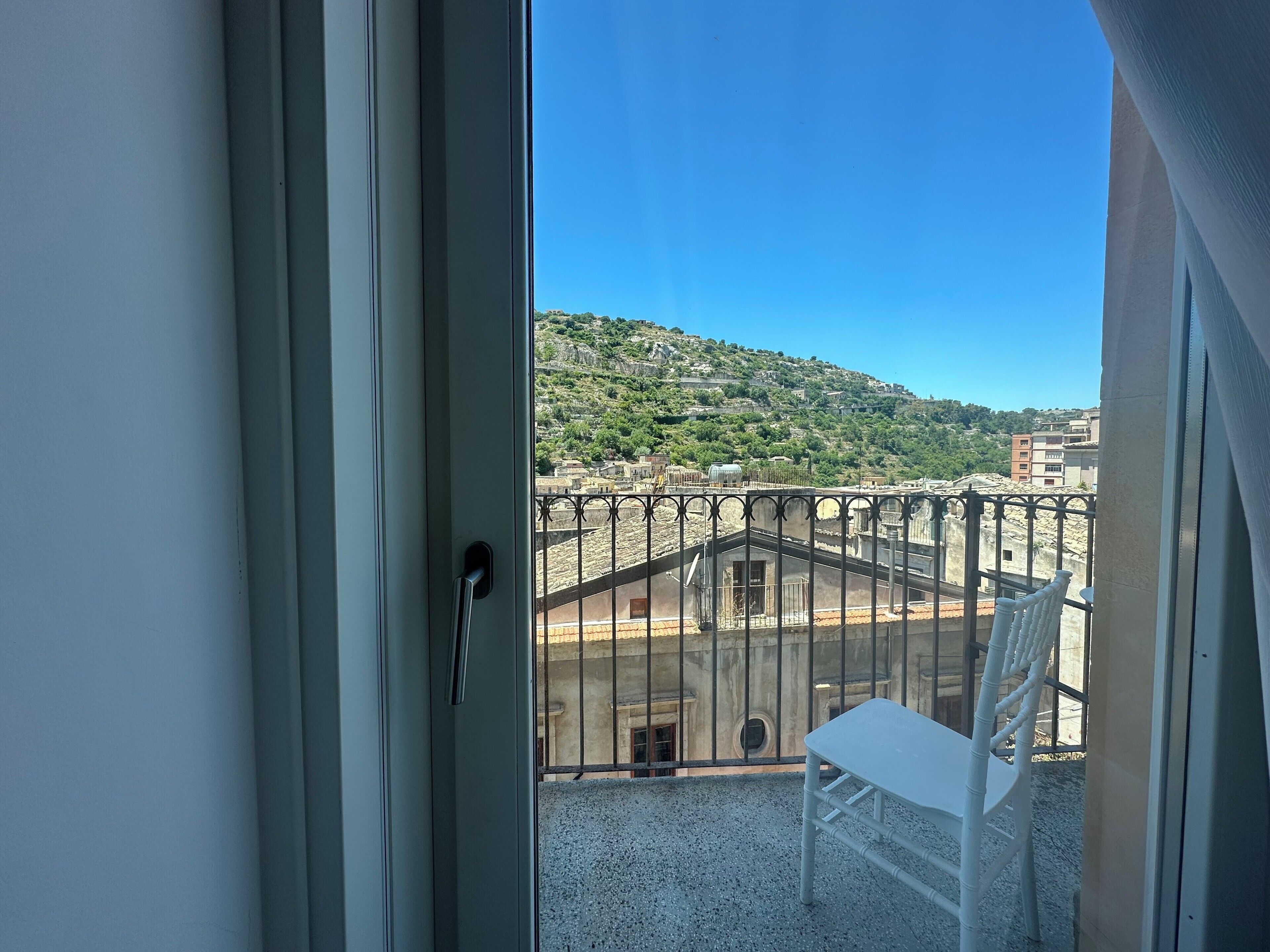 A balcony with a white chair, a metal railing, and a view of a hillside with buildings.