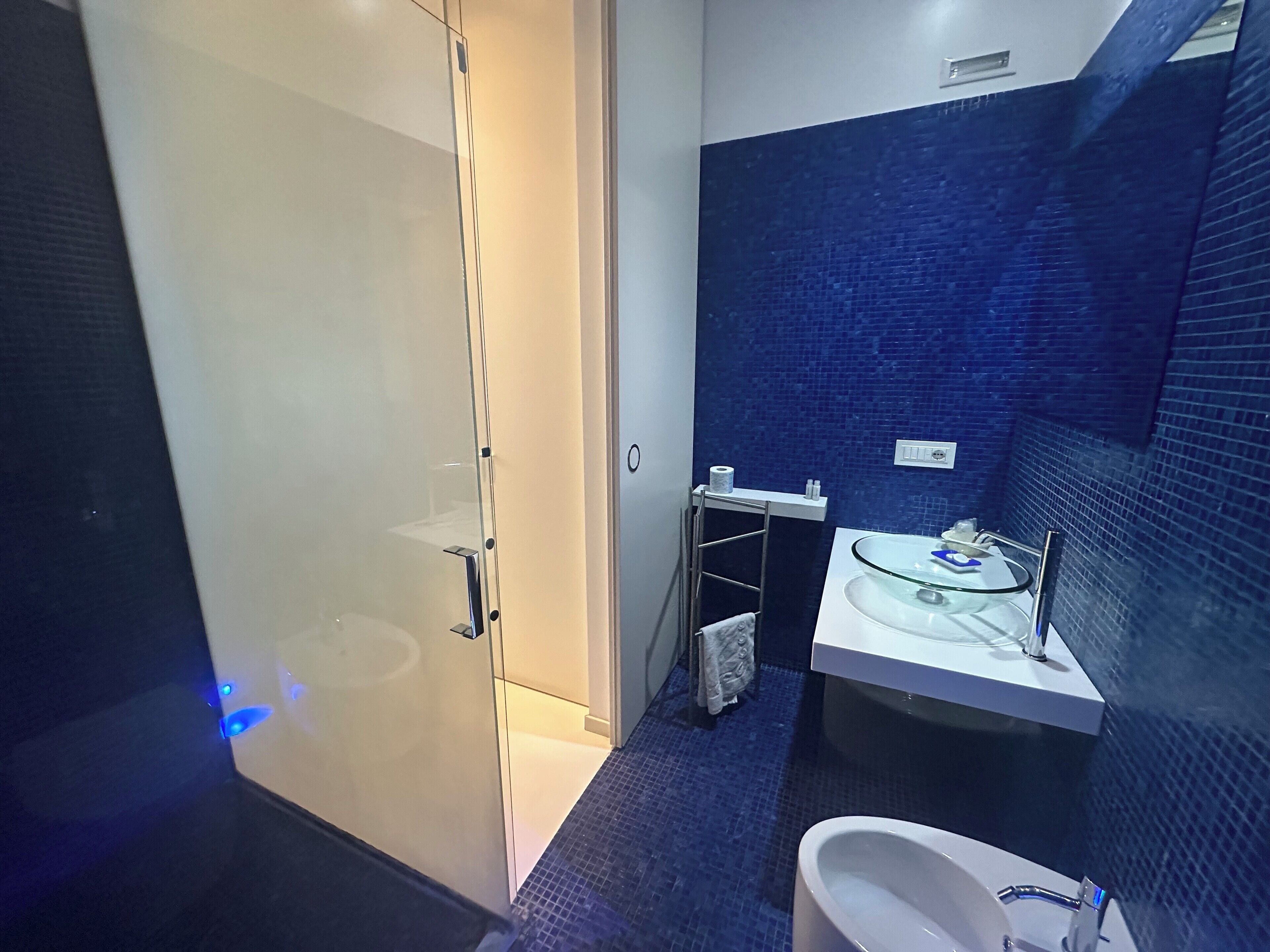A modern bathroom with a glass shower enclosure, a white sink with a glass basin, and a blue-tiled wall.