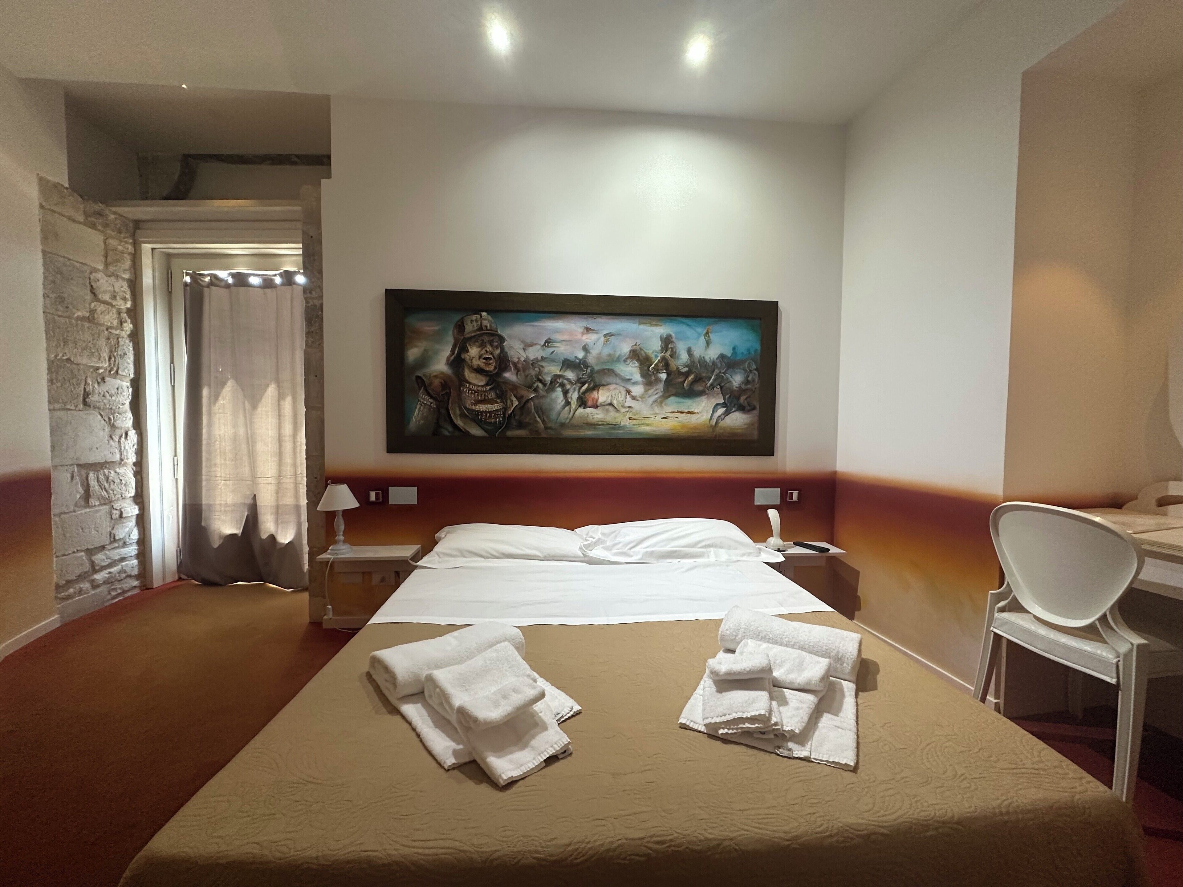 Een hotelkamer met een groot bed, twee witte handdoeken, een schilderij aan de muur, een bureau en een stoel.