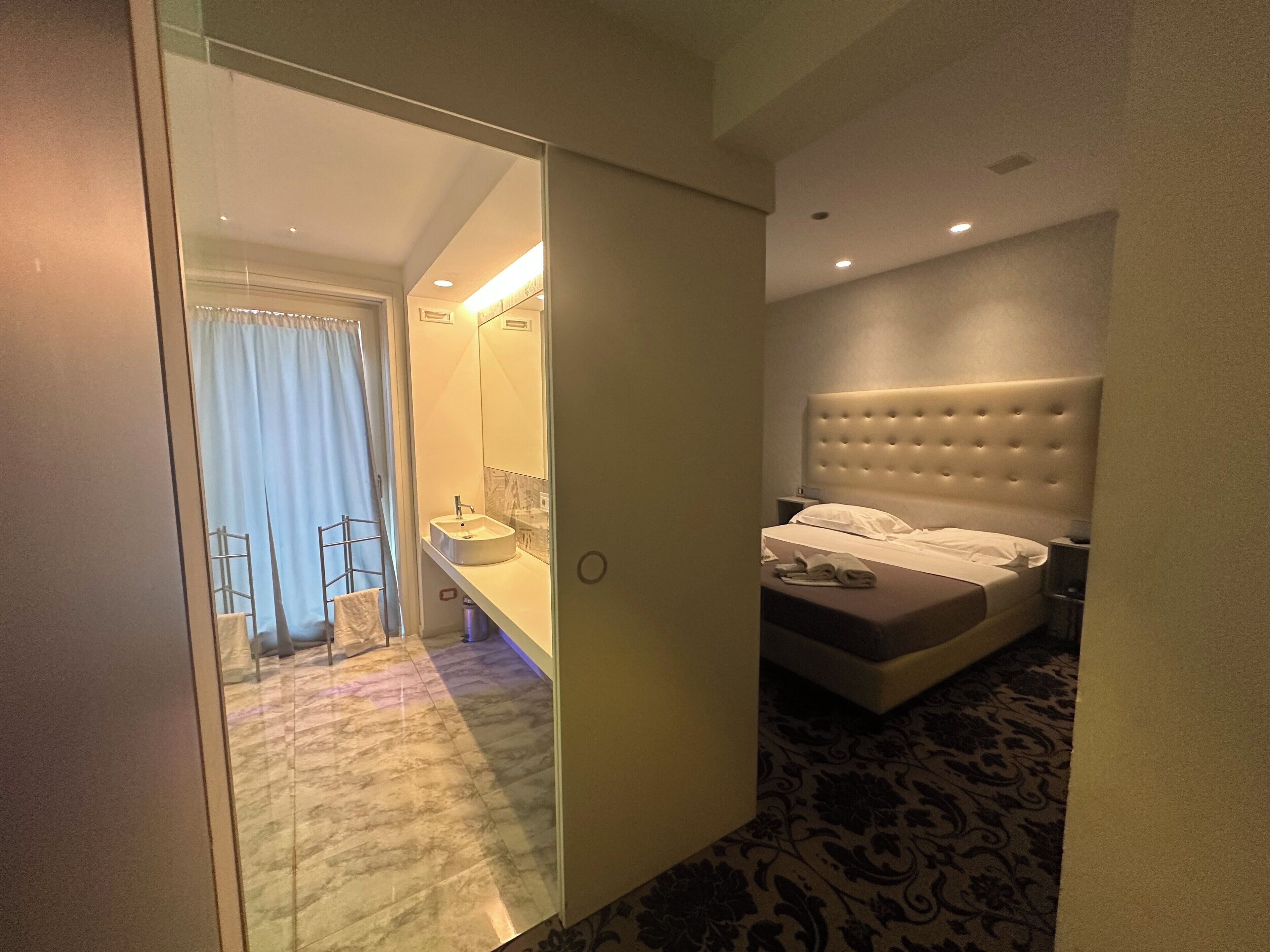A hotel room with a bed, a bedside table, a mirror, and a bathroom visible through an open door.