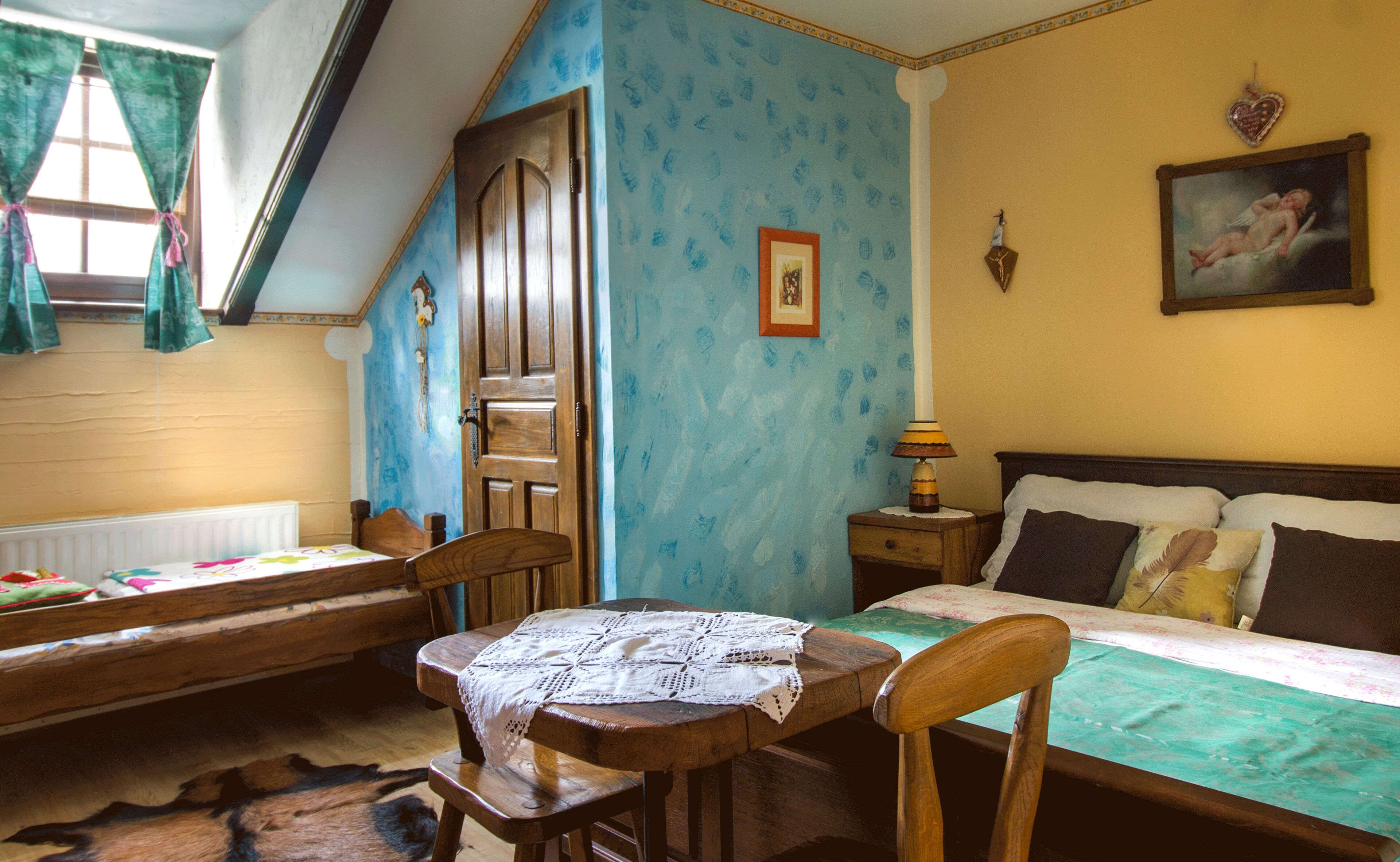A room with a wooden bed, a wooden table with a lace tablecloth, a wooden chair, a nightstand with a lamp, and a painting on the wall.