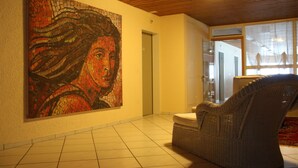 Een hal met een groot mozaïekkunstwerk aan de muur, een rieten stoel en een betegelde vloer.