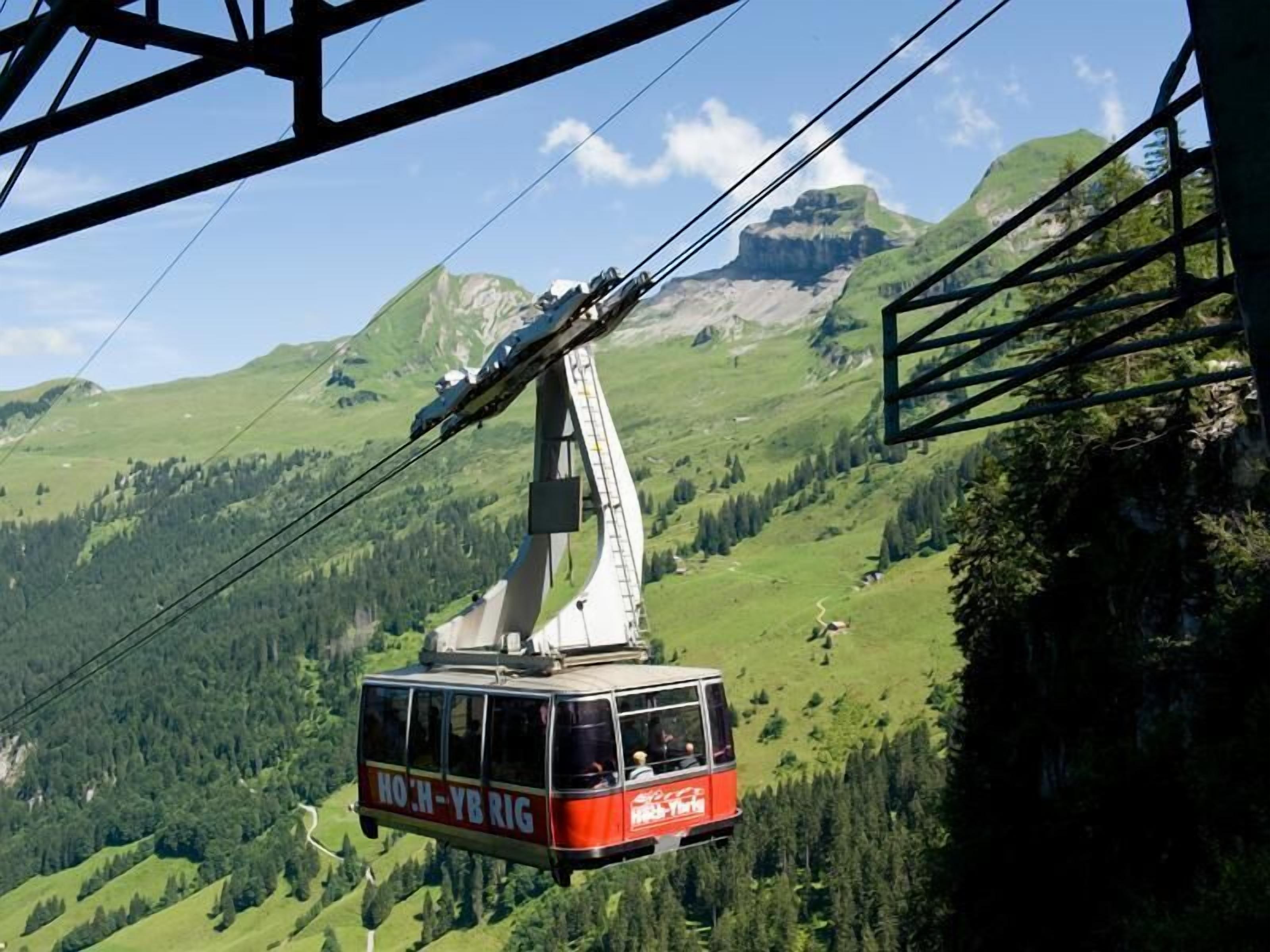 Un teleférico con la marca "HOH-YBRIG", suspendido por cables con montañas al fondo.