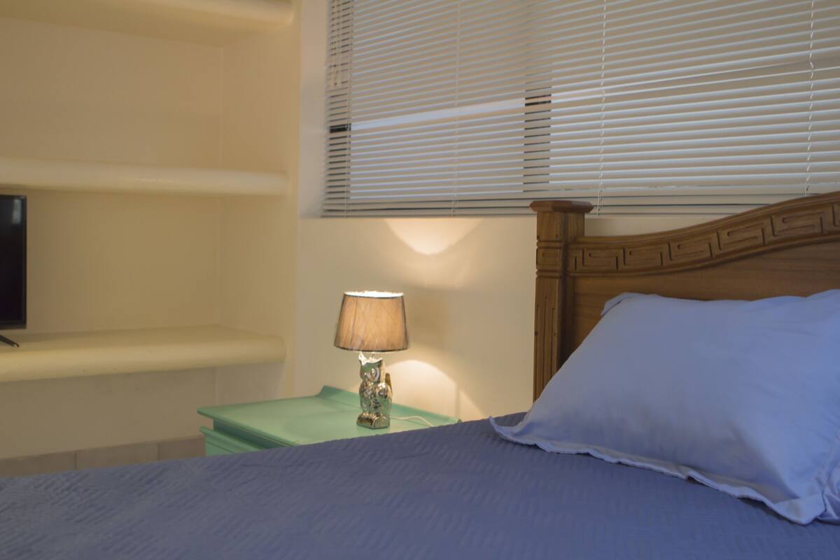 Een slaapkamer met een bed, een nachtkastje met een lamp en een raam met zonwering.