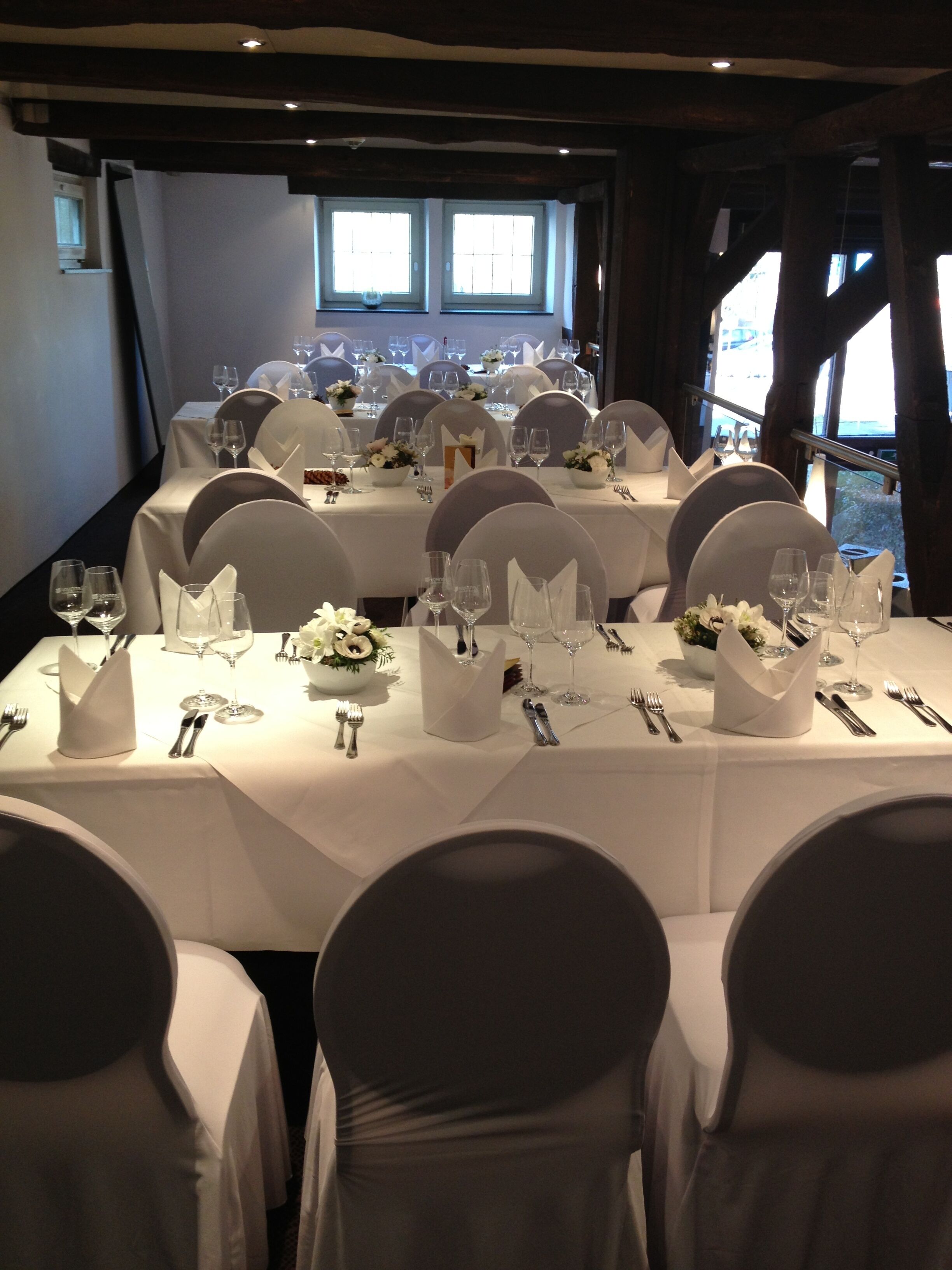 A formal dining room with multiple tables set for a meal, featuring white tablecloths, chairs with white covers, and neatly arranged table settings including wine glasses, plates, and cutlery.