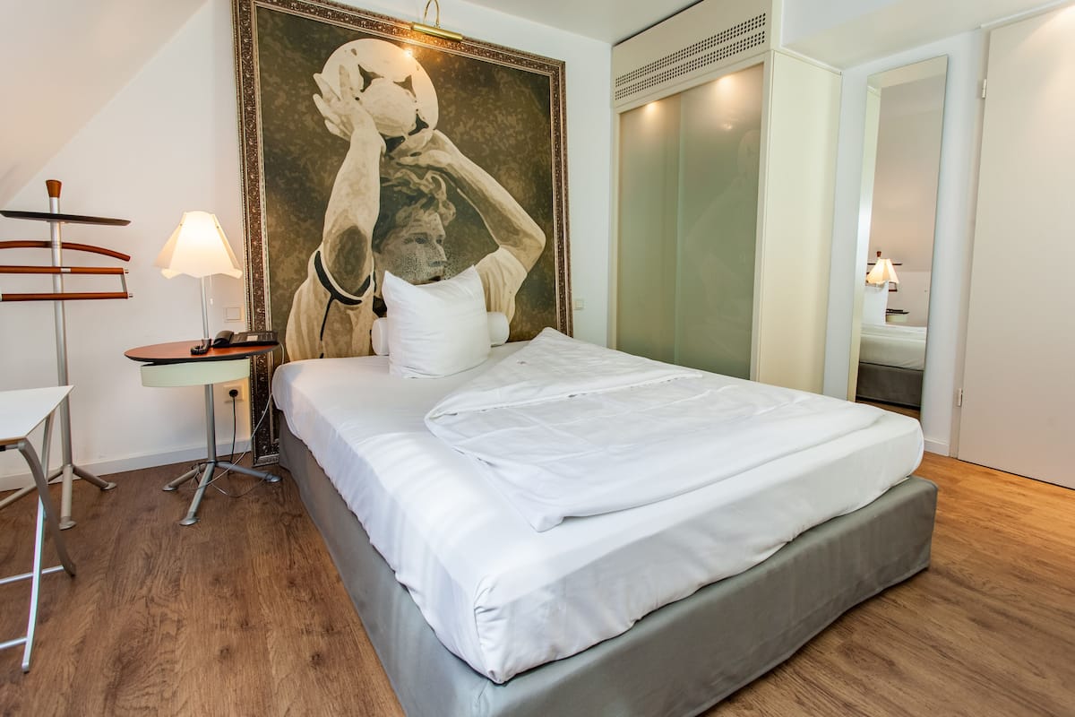 A neatly made bed with white linens, a bedside table with a lamp, and a large abstract painting on the wall.
