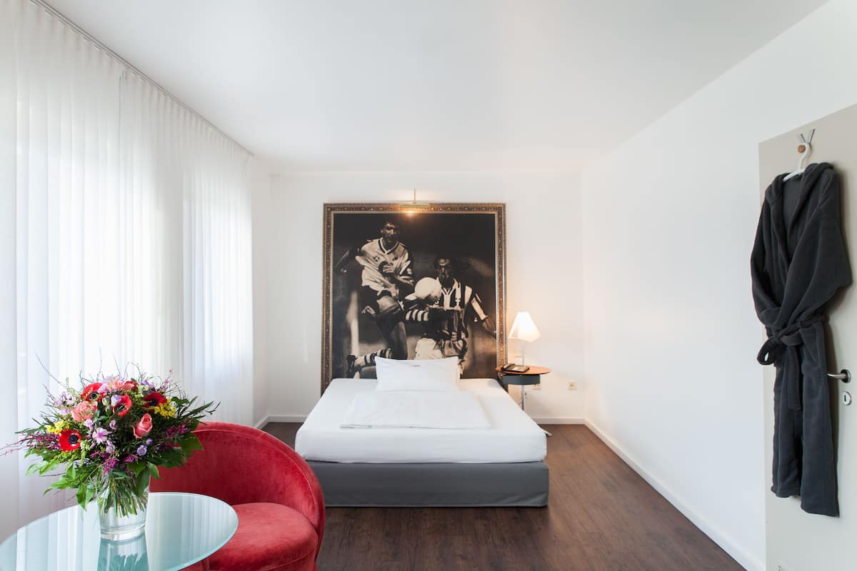 A hotel room with a bed, a red chair, a glass table with a vase of flowers, a framed picture on the wall, and a robe hanging on the door.