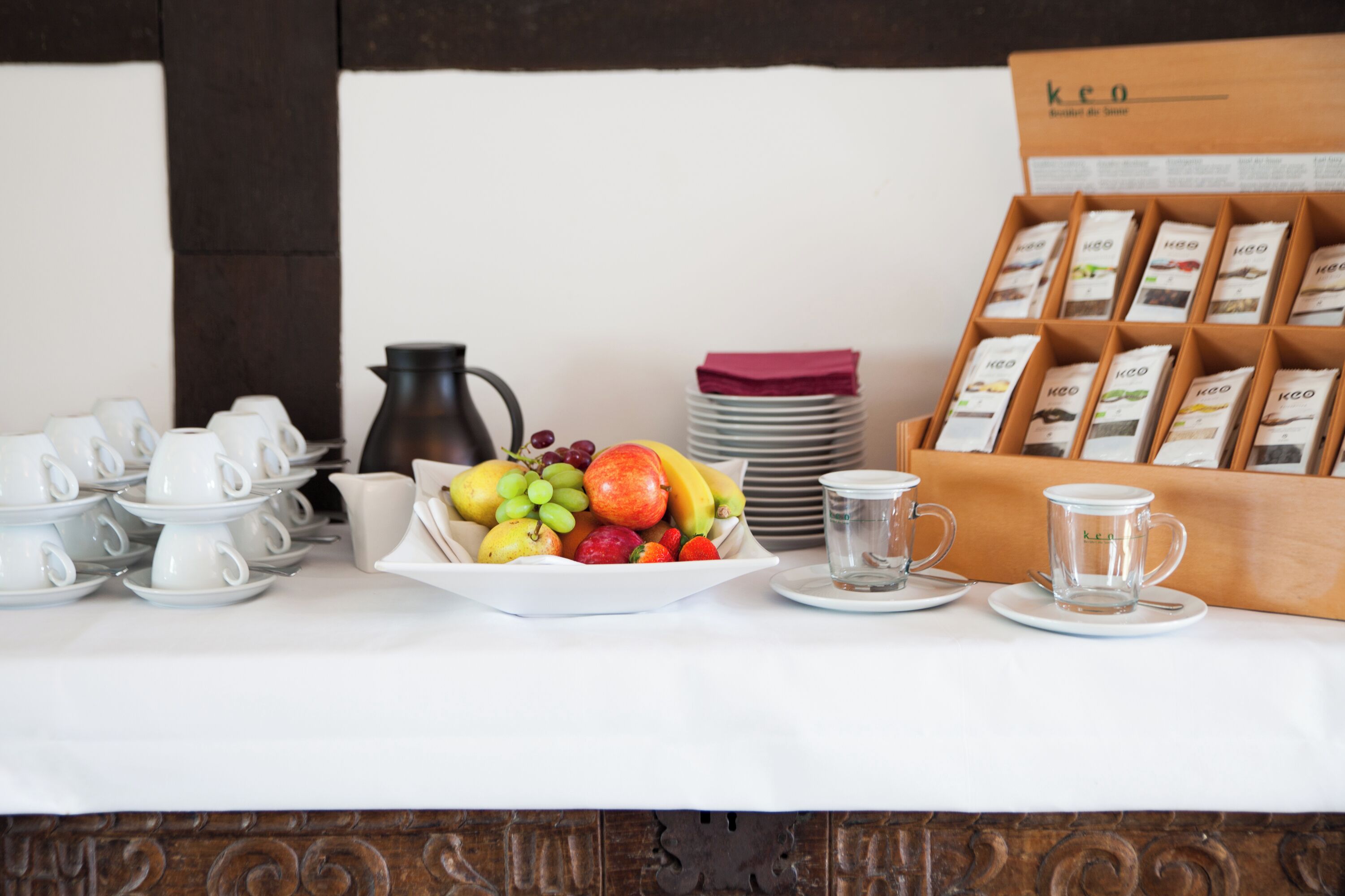 A breakfast setup with white cups, a fruit bowl, a coffee pot, and a box of KEO coffee samples.