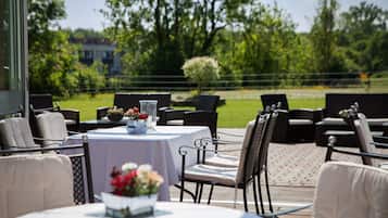 An outdoor dining area with round tables and chairs, surrounded by greenery and a fence.