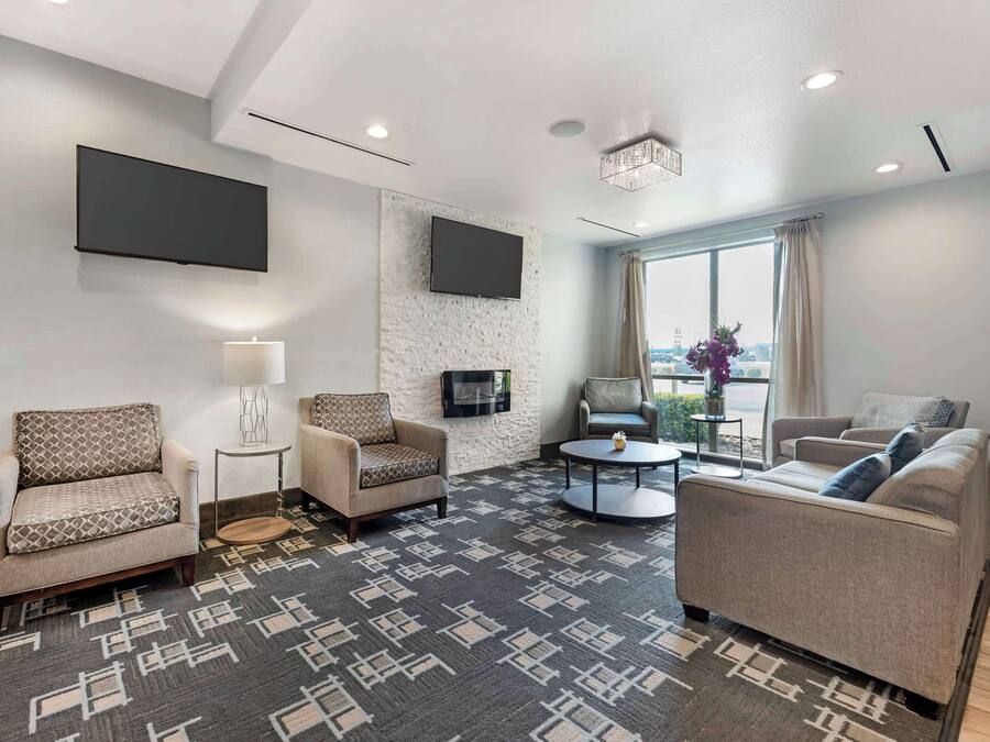 A modern hotel room with a large flat-screen TV mounted on the wall, a comfortable seating area with two sofas and a chair, a small round coffee table, and a patterned rug.