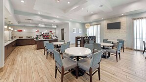 Een moderne hotel eetruimte met ronde tafels en stoelen, een keuken op de achtergrond en een flatscreen televisie aan de muur.