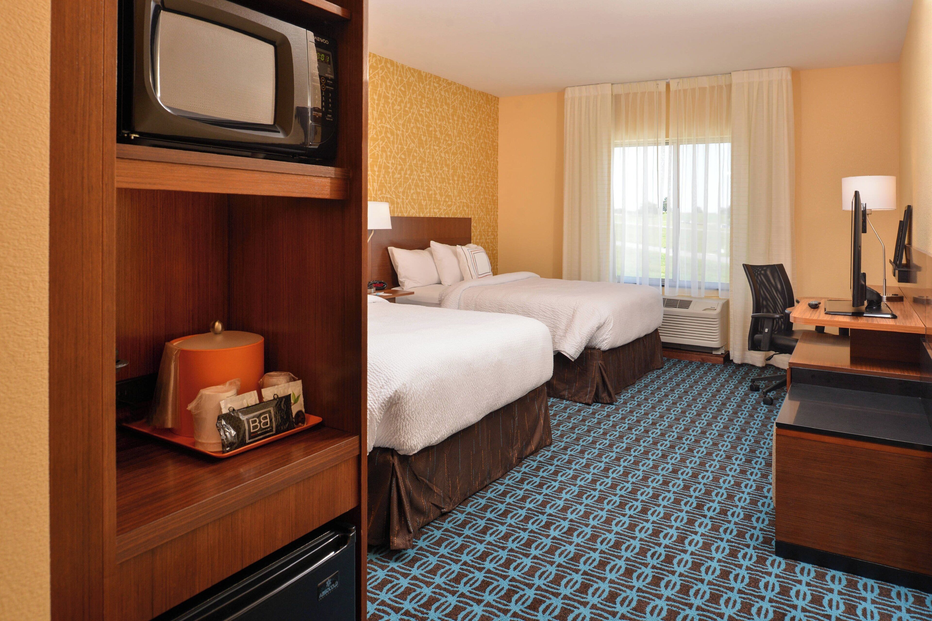 A hotel room with two beds, a microwave, a coffee maker, a desk with a computer, and a window with curtains.