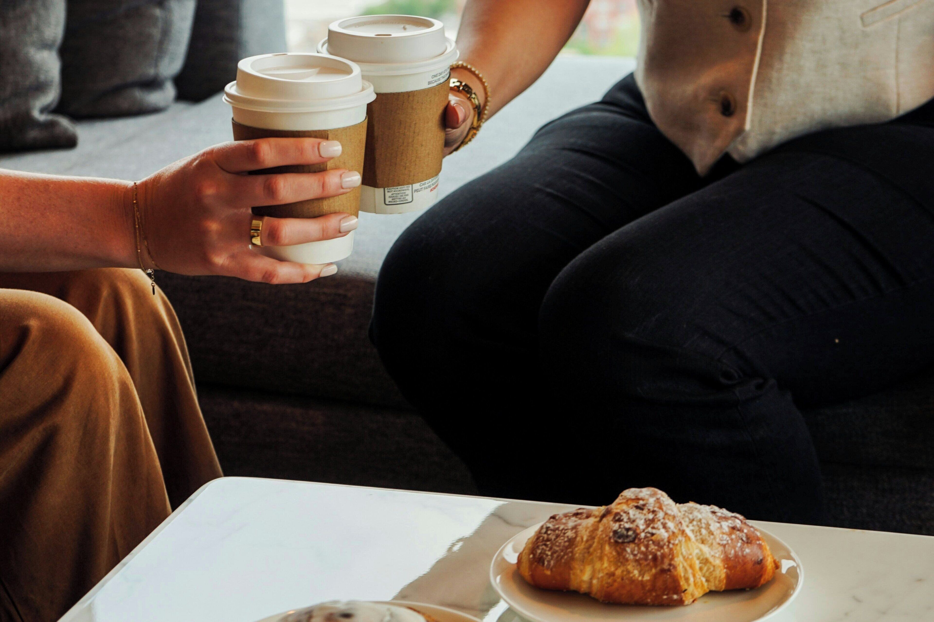 A person holding two coffee cups and a plate with a pastry.