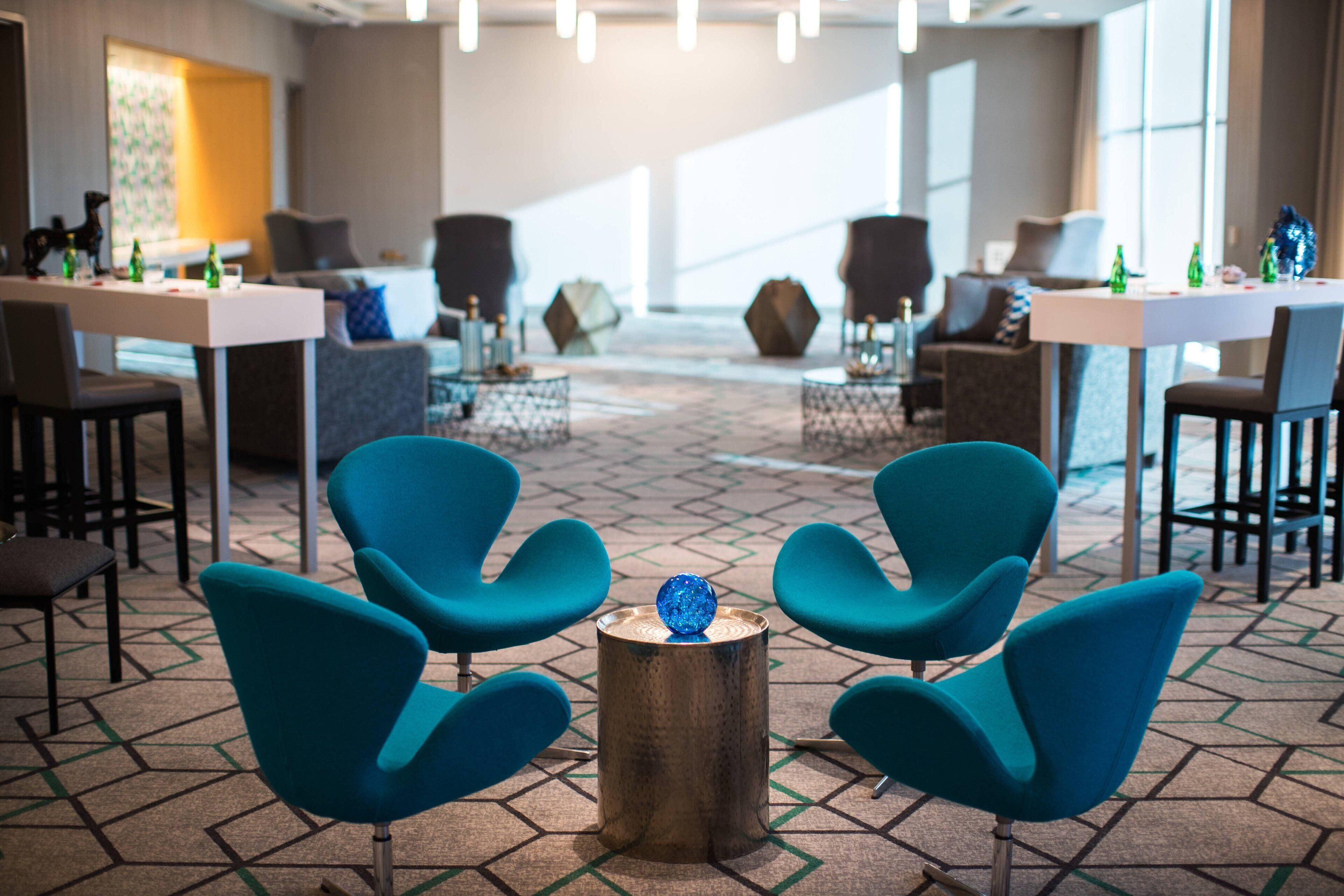 Un moderno vestíbulo de hotel con sillas azul verdosas, una mesa redonda de madera y un adorno decorativo azul, sobre una alfombra con motivos geométricos.