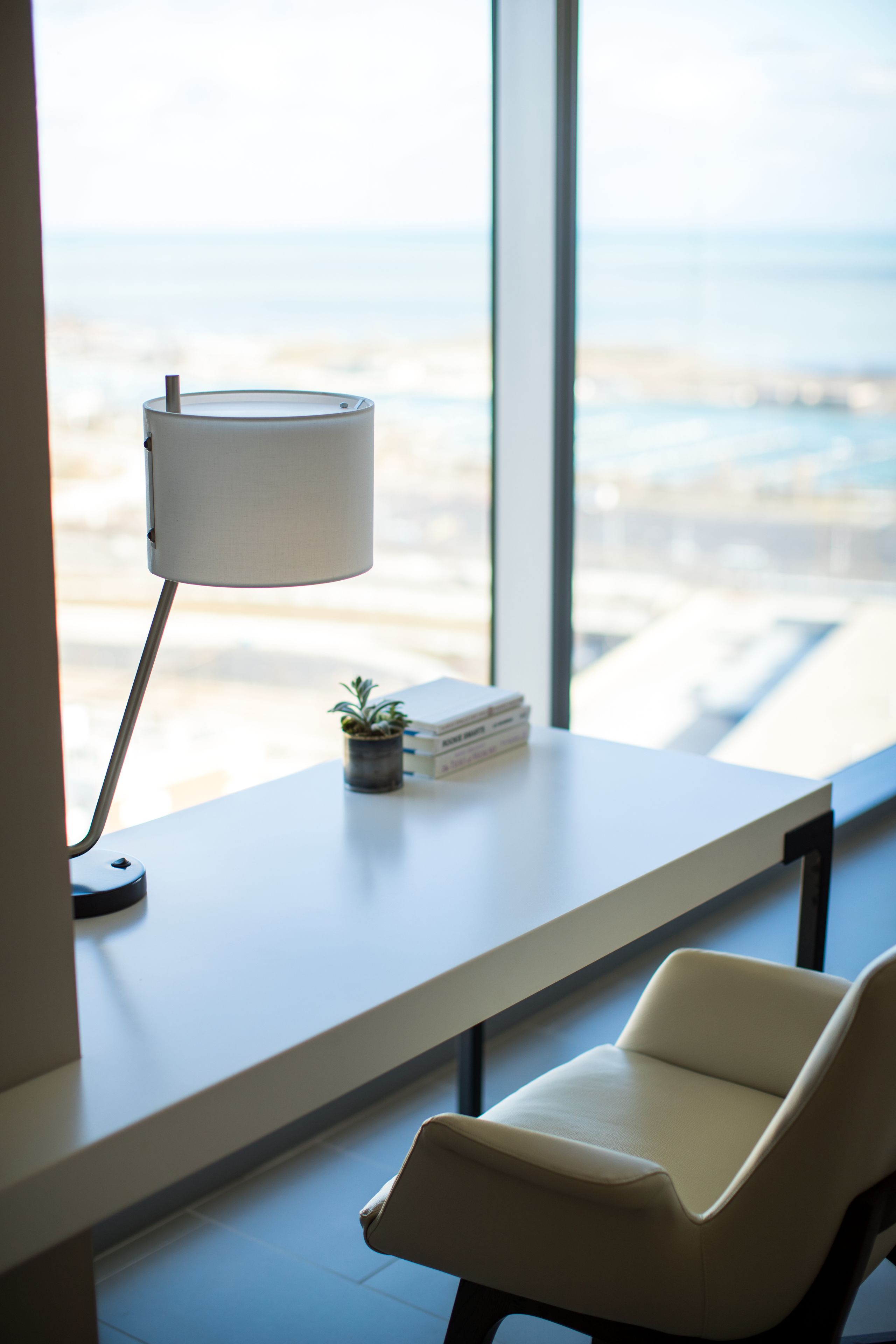 Ein modernes Zimmer mit großem Fenster, Schreibtisch mit Lampe, Stuhl und einer kleinen Pflanze.