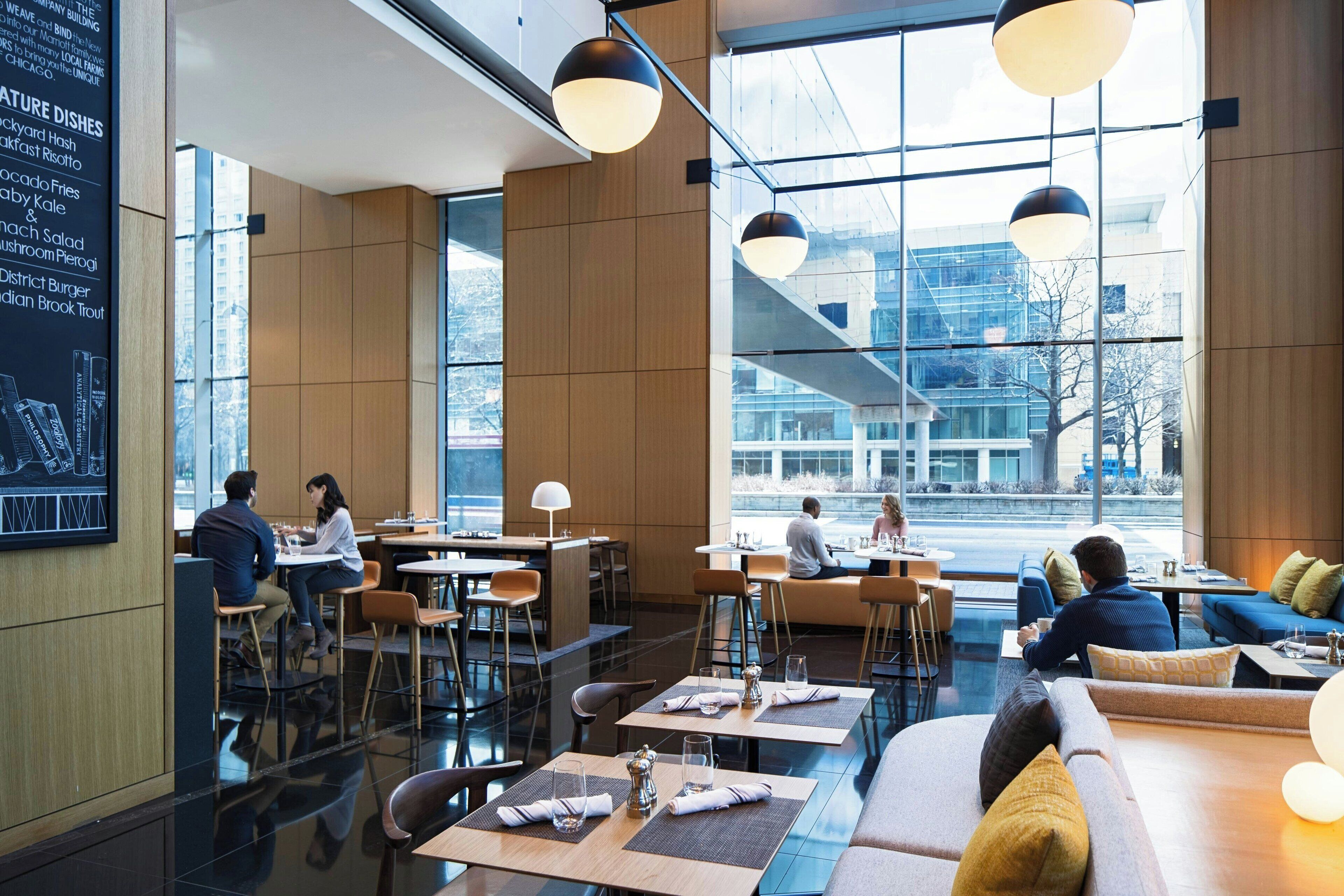 Ein modernes Restaurant mit hohen Decken, großen Fenstern und einer Mischung aus Sitzbereichen, darunter Tische und Sofas.