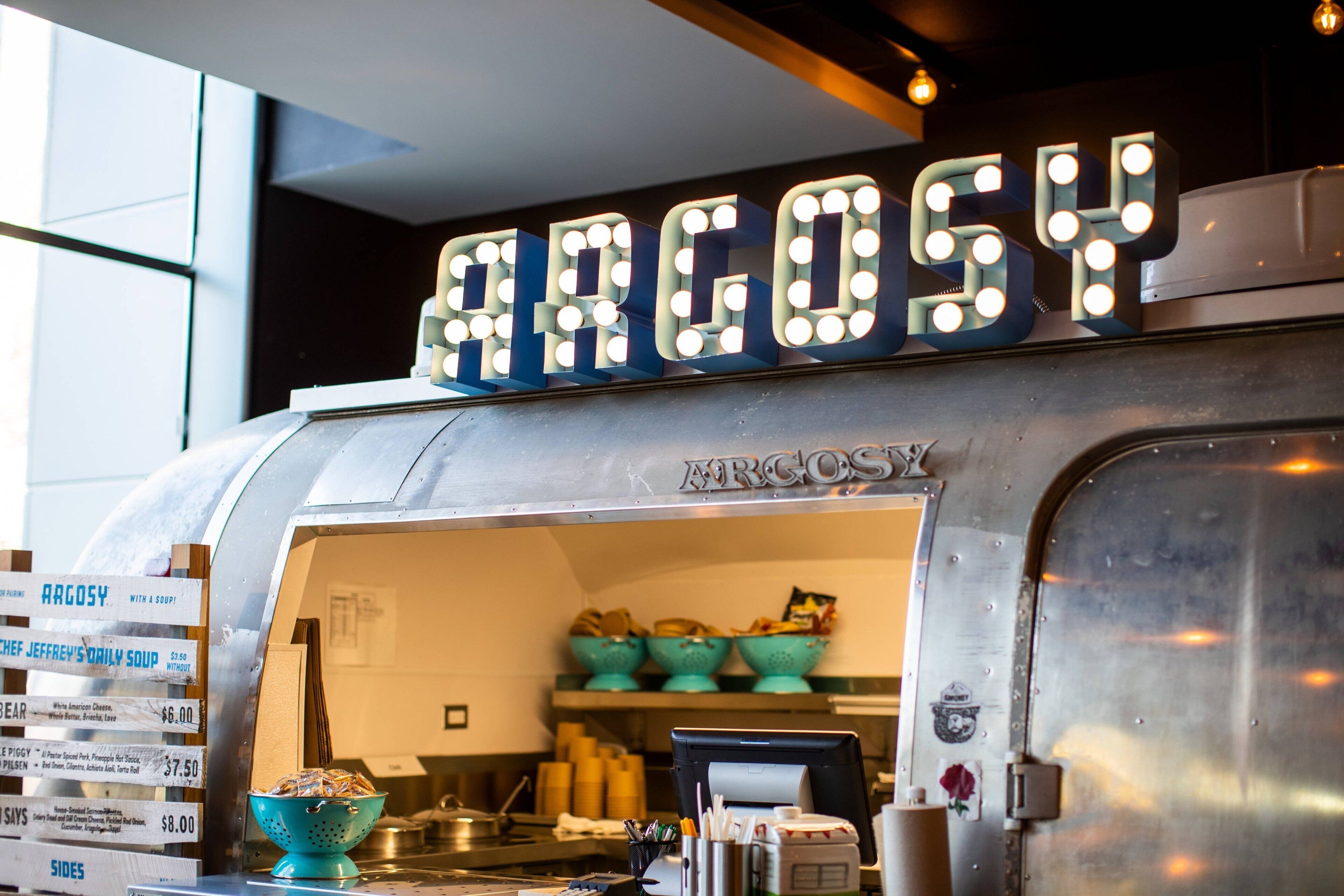 A food truck with a sign that reads "ARGOSY" and a menu board listing items like "CHEF JEFFREW'S DAILY SOUP" and "BEAR PIGG".