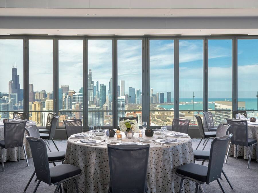 Ein Essbereich mit runden Tischen, Stühlen und Blick auf die Stadt durch große Fenster.