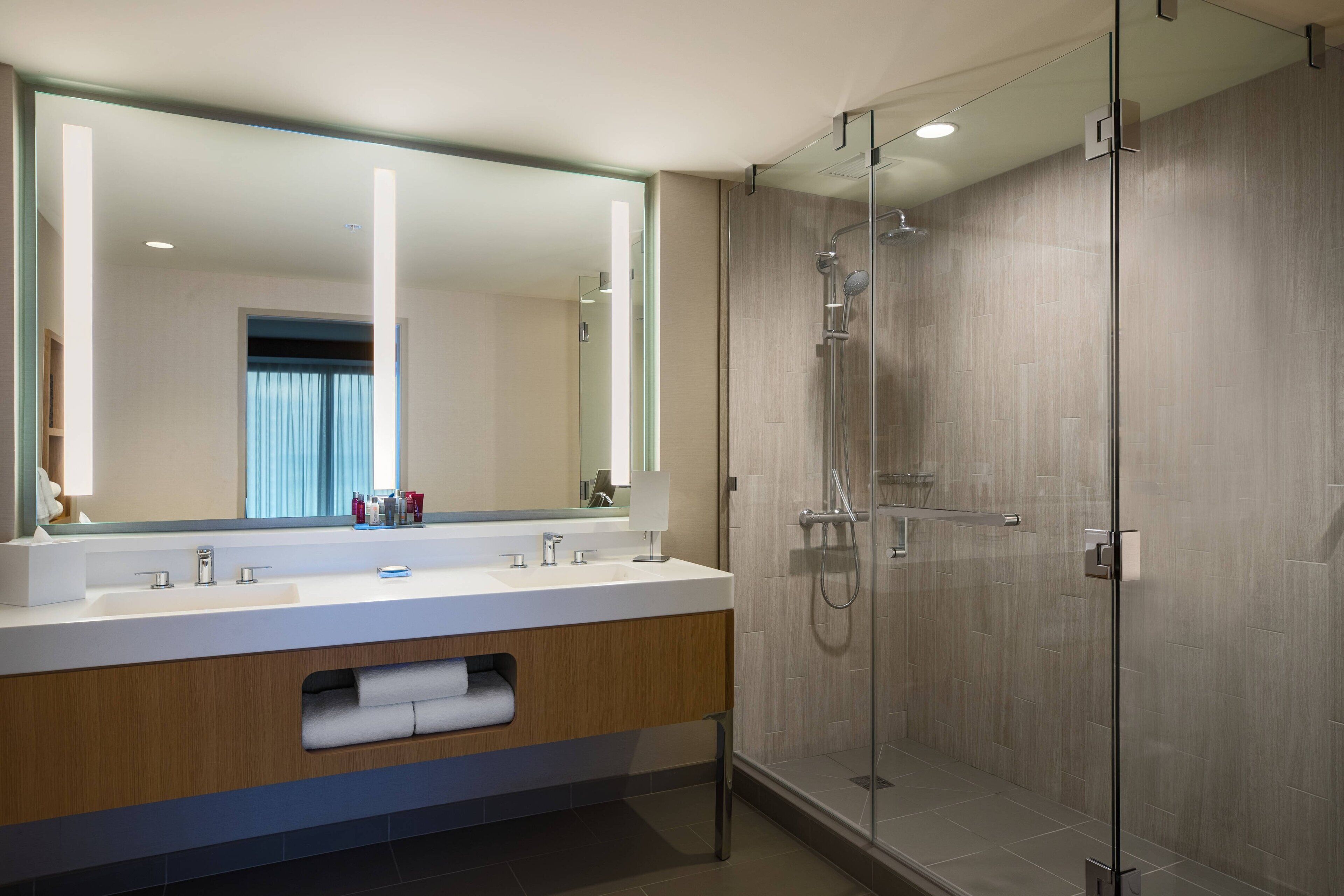 Un baño moderno con un espejo grande, lavamanos dobles, cabina de ducha de vidrio y tocador de madera.
