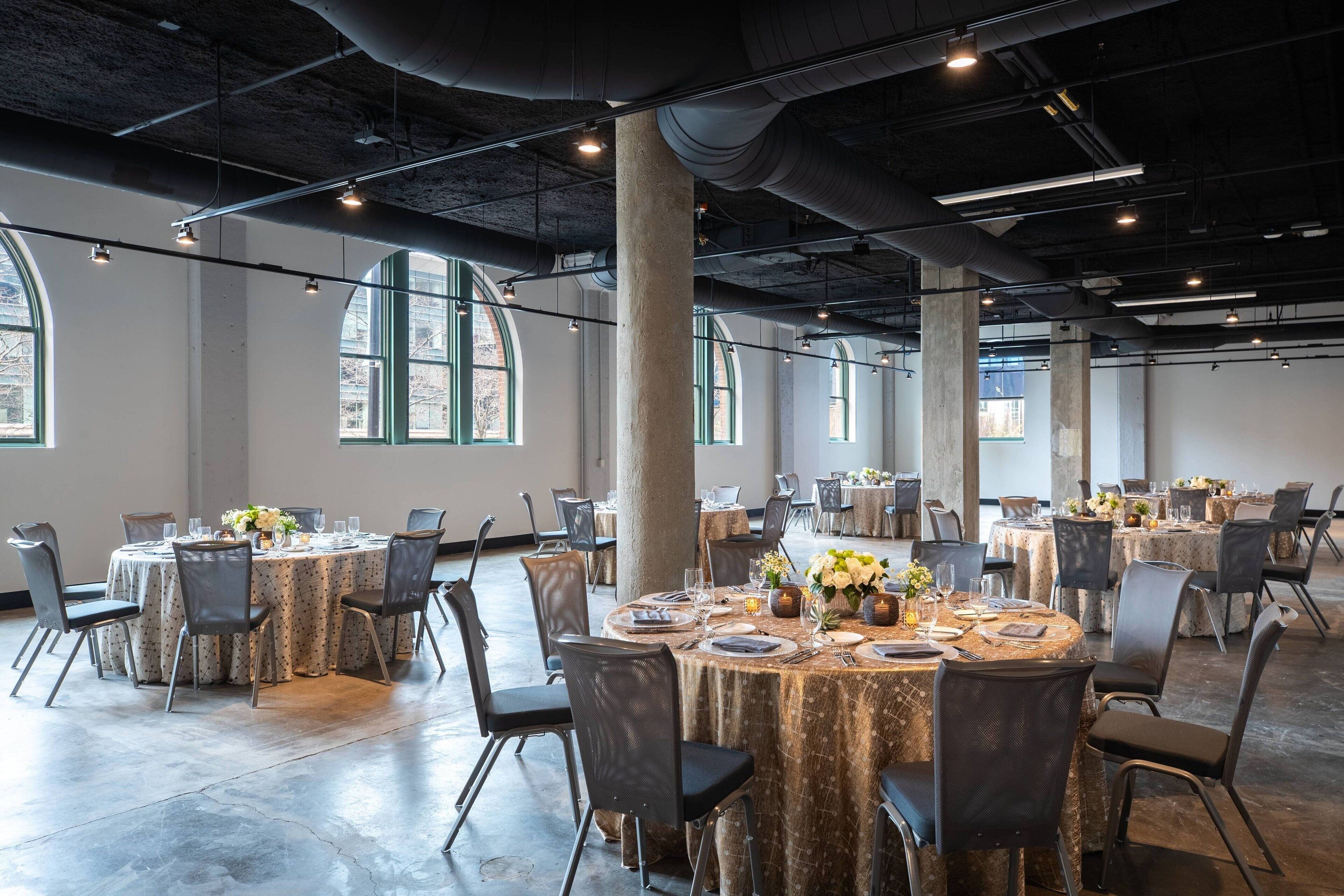 Un vaste hall doté de nombreuses tables rondes disposées pour les repas, de grandes fenêtres et de tuyaux apparents au plafond.