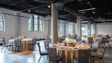 Un vaste hall doté de nombreuses tables rondes disposées pour les repas, de grandes fenêtres et de tuyaux apparents au plafond.