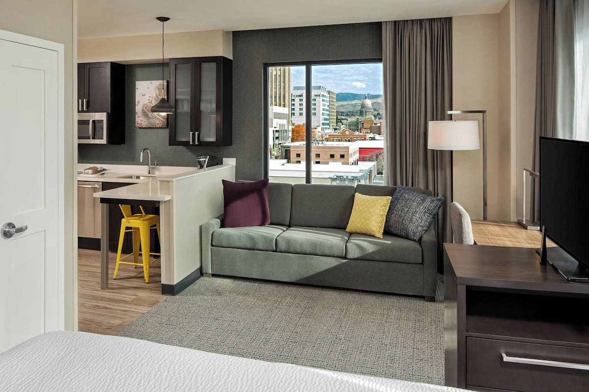 A modern hotel room with a sofa, a TV, a desk with a chair, a large window with a city view, and a lamp.