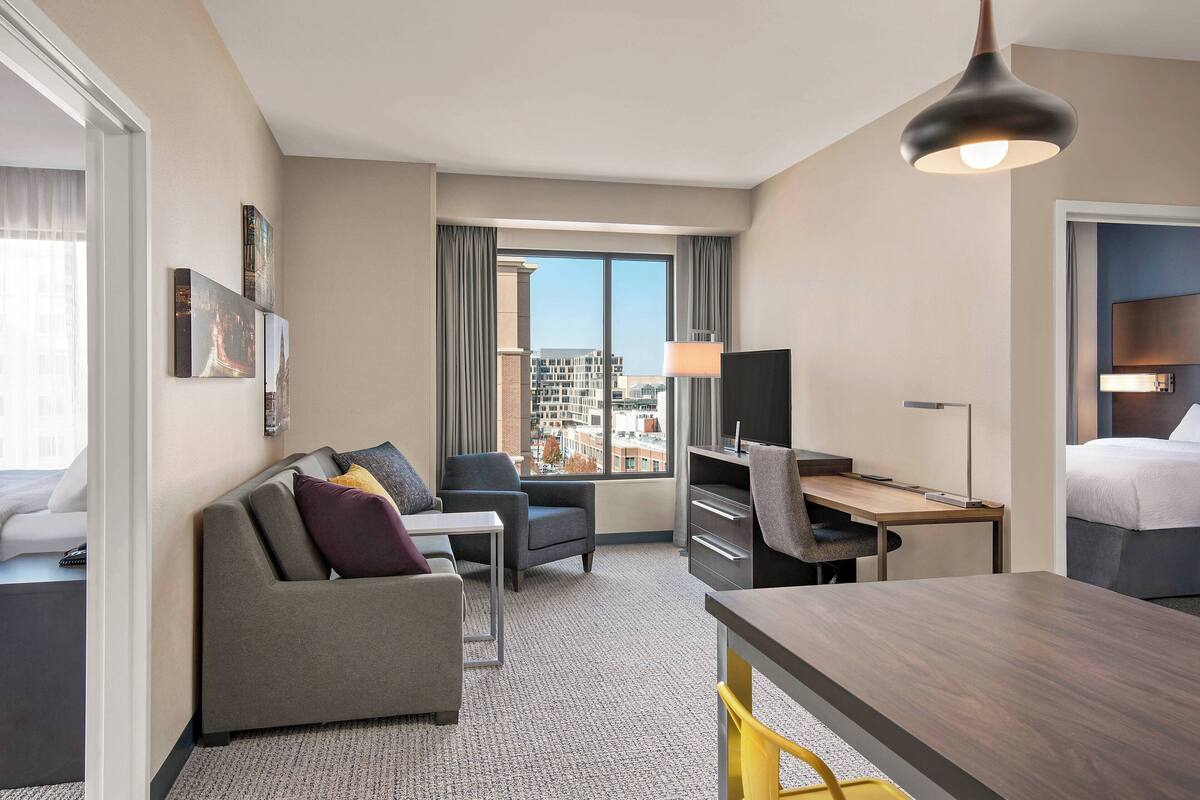A hotel room with a sofa, a desk, a chair, a bed, a TV, and a window with a city view.