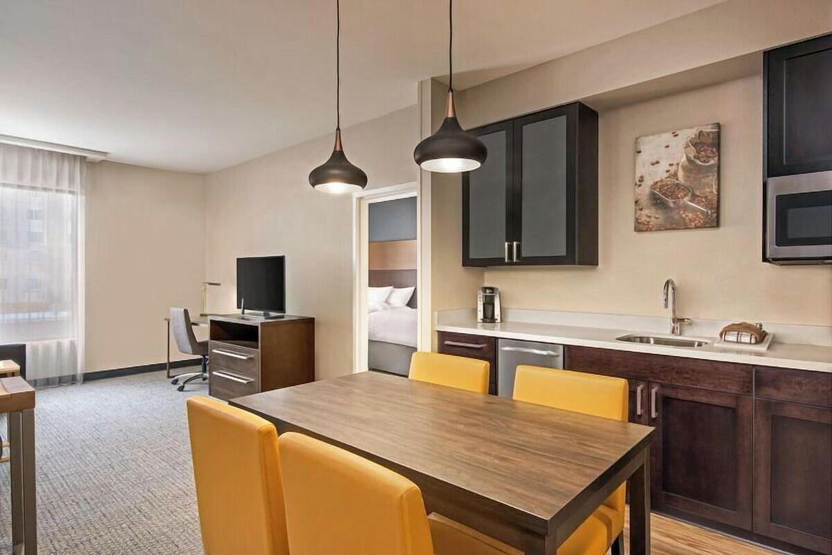 A hotel room with a wooden dining table, yellow chairs, a TV, a desk, a bed, a kitchenette, and a painting on the wall.