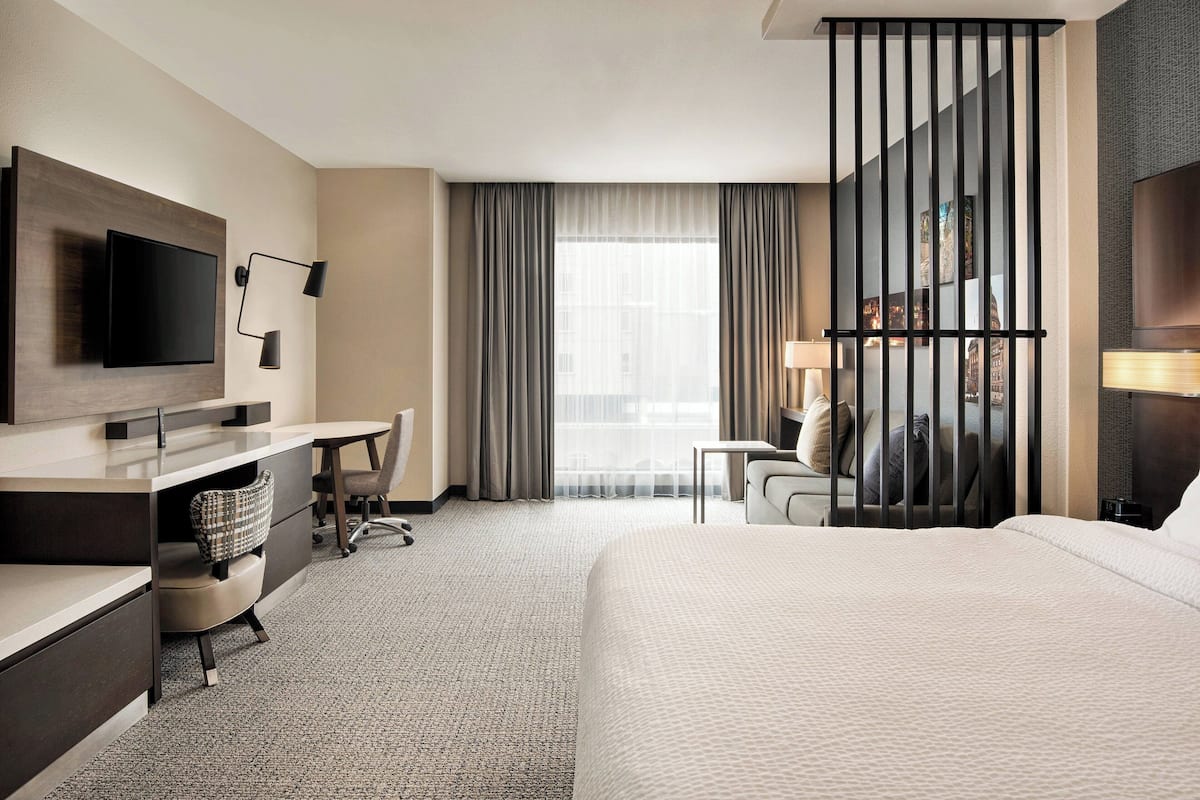 A hotel room with a bed, a desk with a chair, a TV, and a large window with curtains.