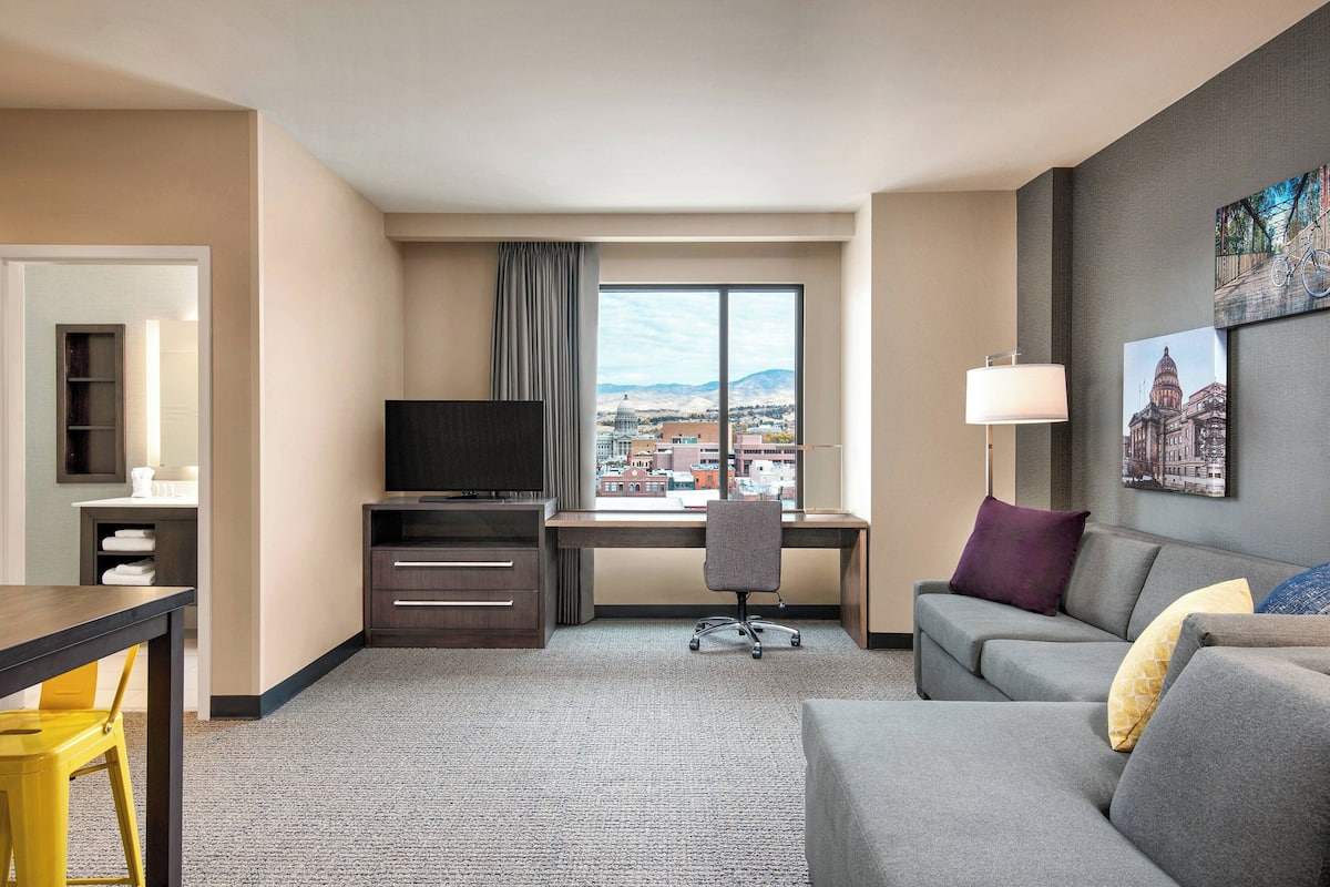A hotel room with a desk, a television, a sofa, and a large window offering a city view.