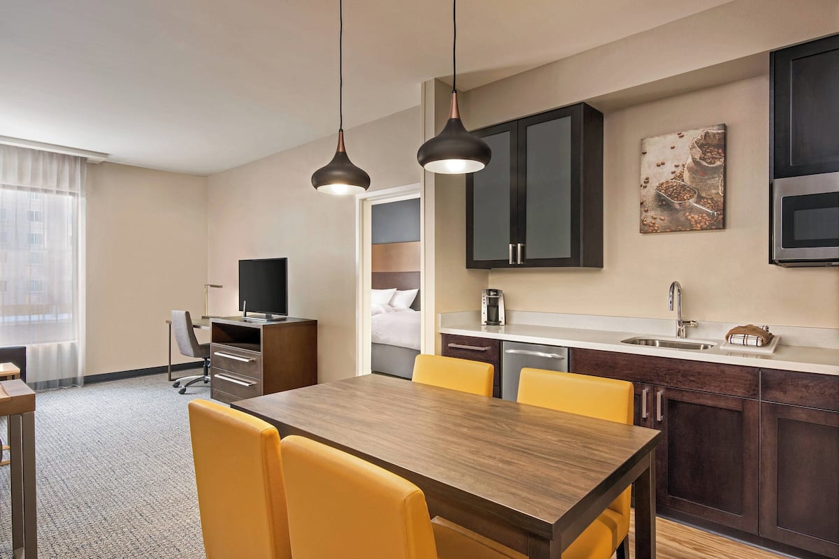 A hotel room with a wooden dining table, yellow chairs, a kitchen area with dark cabinets, a coffee maker, and a television.