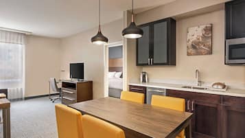A hotel room with a wooden dining table, yellow chairs, a kitchen area with dark cabinets, a coffee maker, and a television.