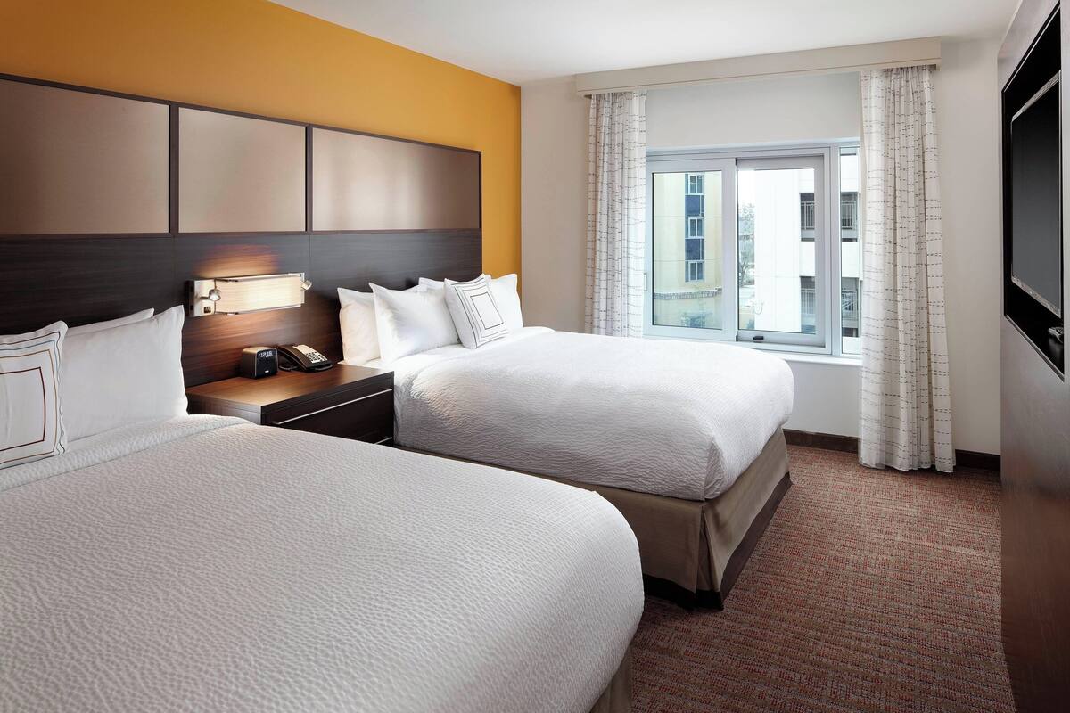 A hotel room with two beds, a nightstand, a lamp, a window with curtains, and a view of buildings outside.