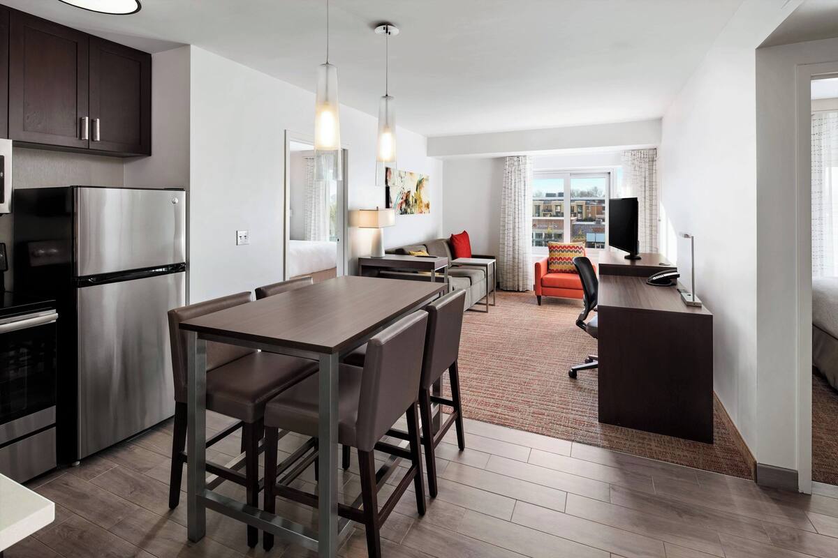 A modern kitchen with a stainless steel refrigerator, a dining area with a wooden table and chairs, a small seating area with a sofa and a TV, and a view of the city through large windows.
