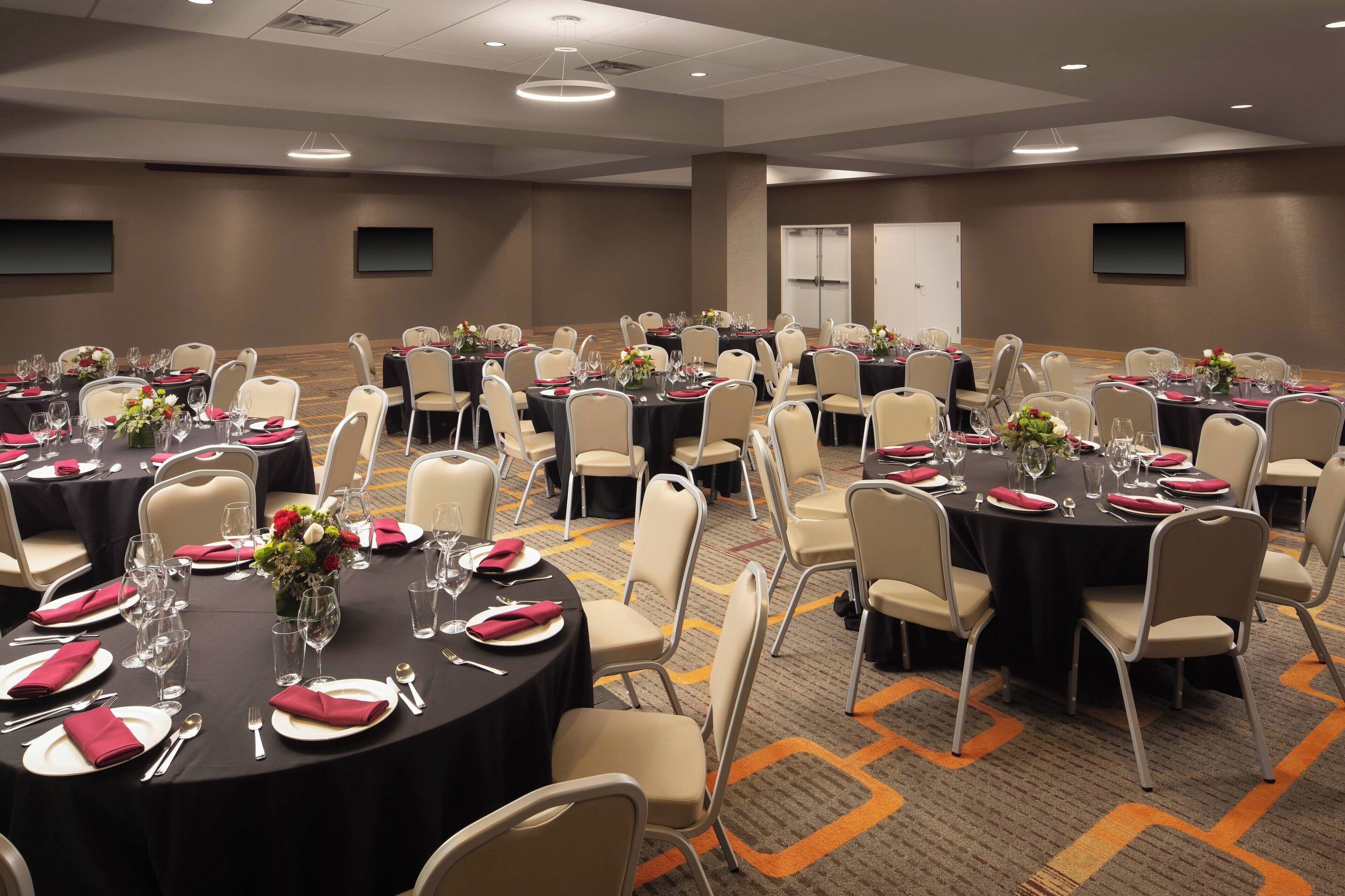 Un salón de conferencias con varias mesas redondas, cada una con platos blancos, servilletas rojas y centros de mesa con flores.