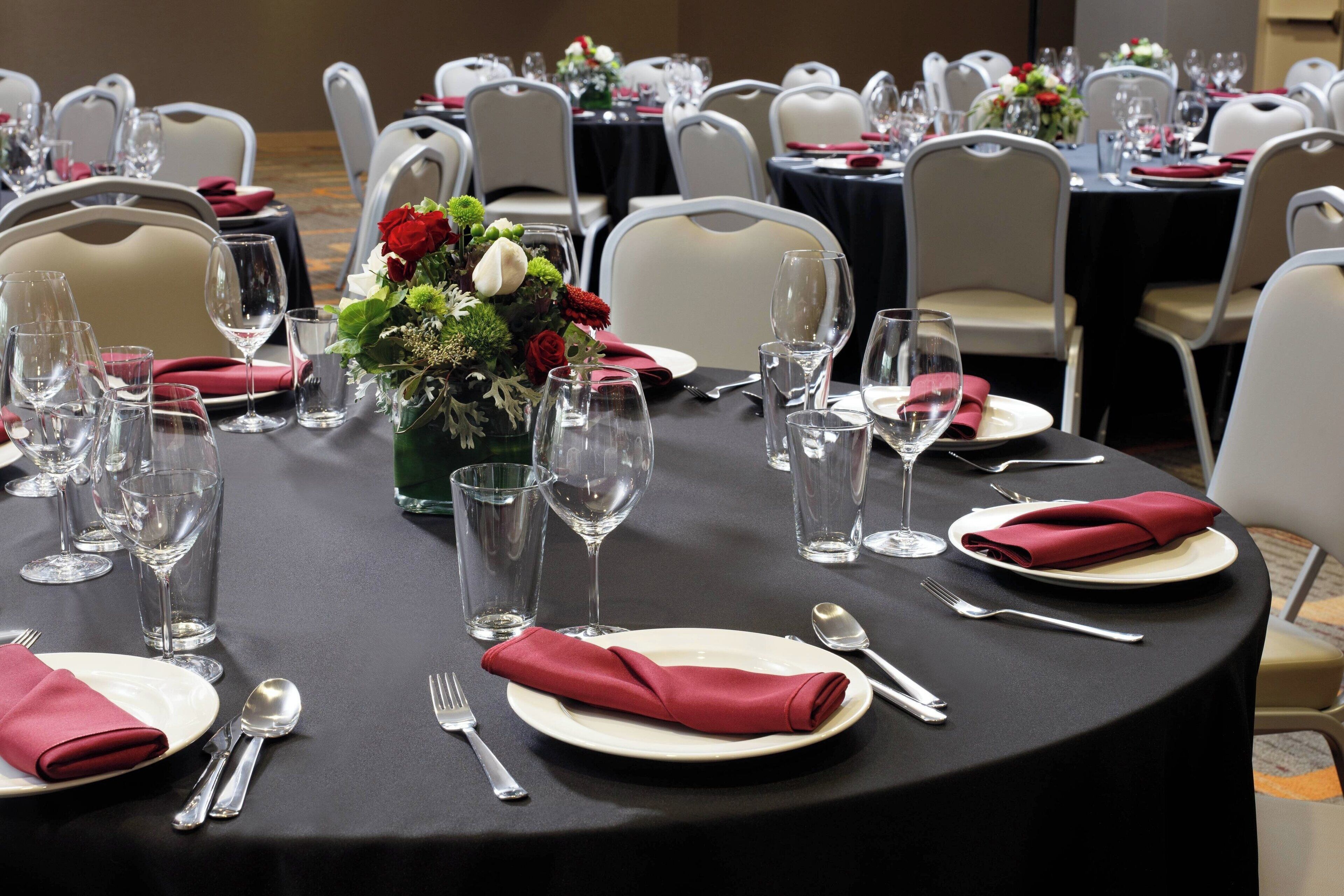 Un salón de banquetes con mesas redondas dispuestas para el servicio de comidas, incluyendo platos, cubiertos y copas de vino.