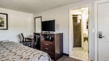 A hotel room with a bed, a desk with a television, a chair, and a wardrobe. There is a bathroom visible through an open door.