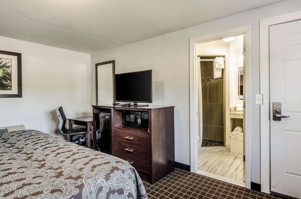 Habitación de hotel con cama, escritorio con televisor, una silla y armario. Se ve un baño a través de una puerta abierta.