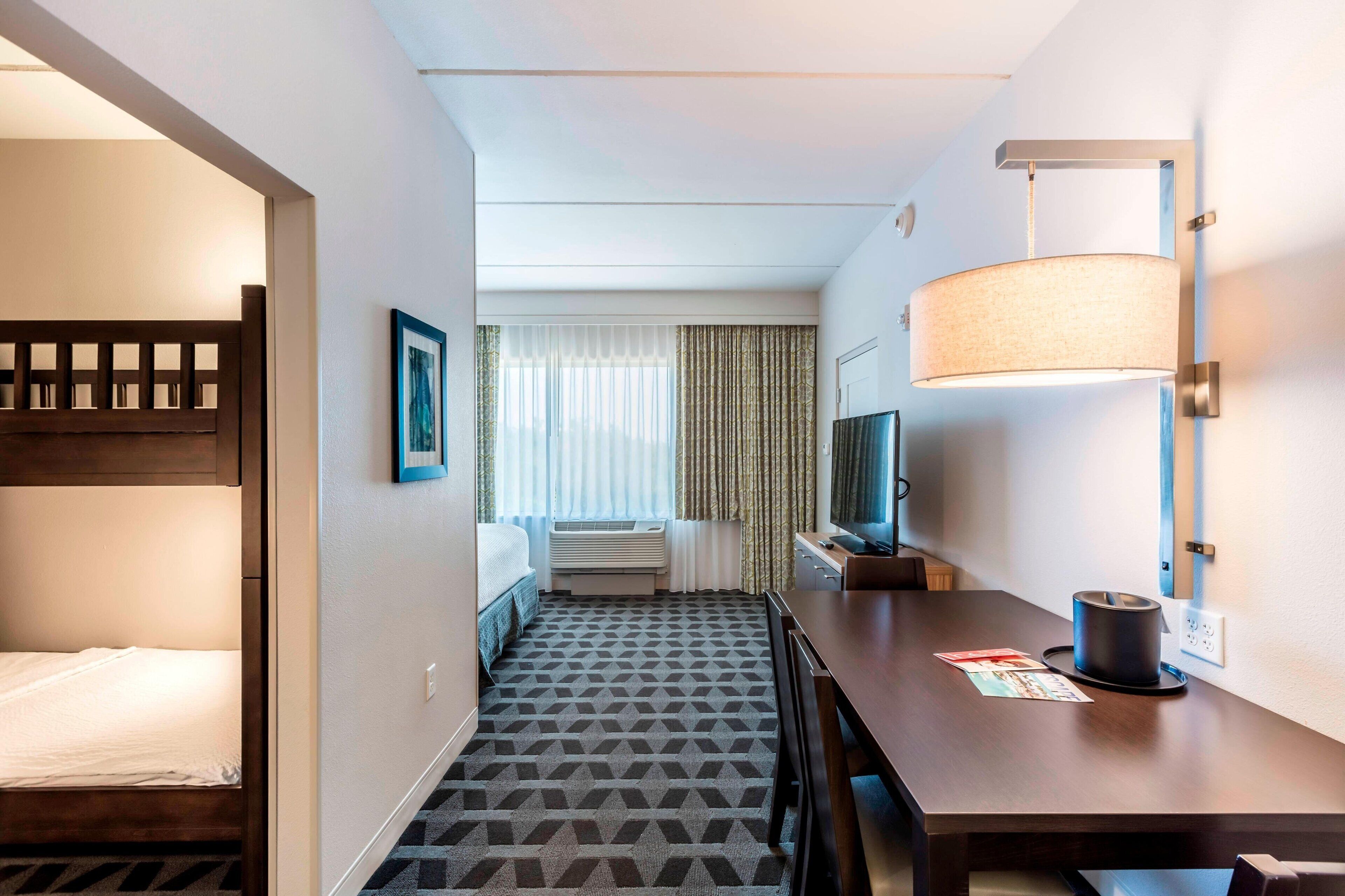 Et hotelværelse med køjeseng, skrivebord med lampe, fjernsyn og et vindue med gardiner.