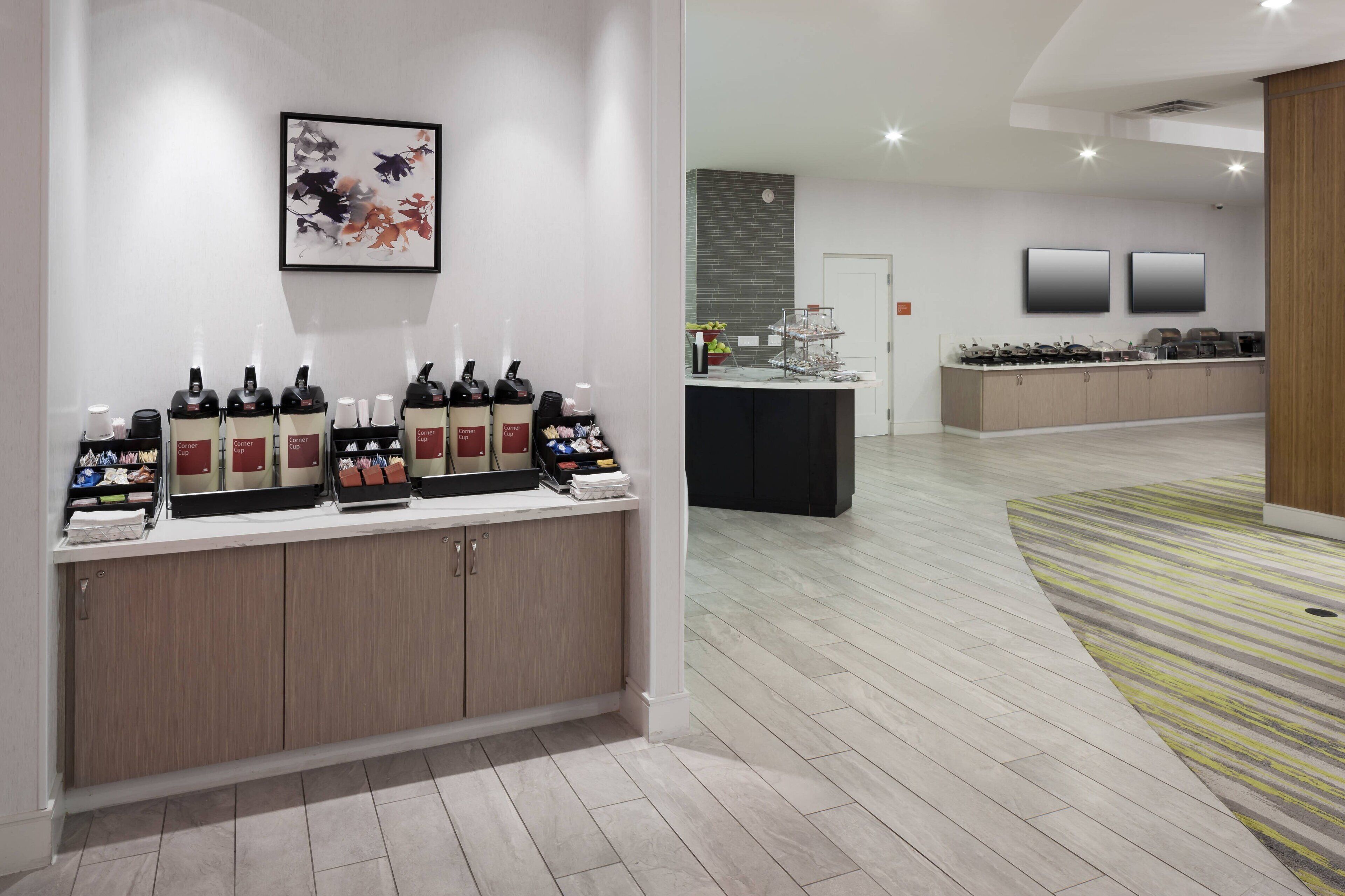 A modern hotel lobby with a reception desk, a coffee station, and a large mirror.
