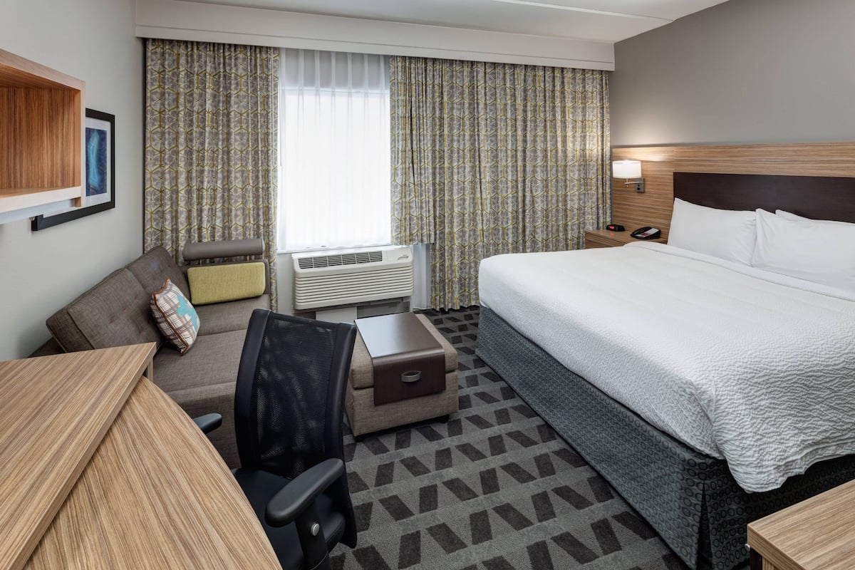 A hotel room with a large bed, a desk, a chair, a small table, an air conditioning unit, and curtains.