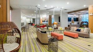 Un moderno vestíbulo de hotel con un área de estar central, una escalera y una cocina al fondo.